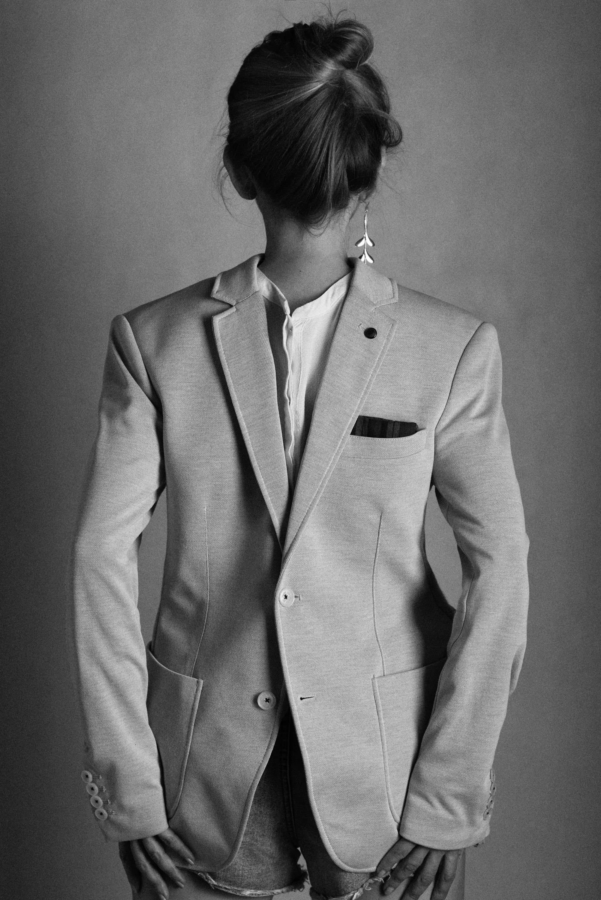Black and white photograph of a person with an updo hairstyle, wearing a tailored blazer, a collared shirt, and distressed denim shorts, standing against a plain background.