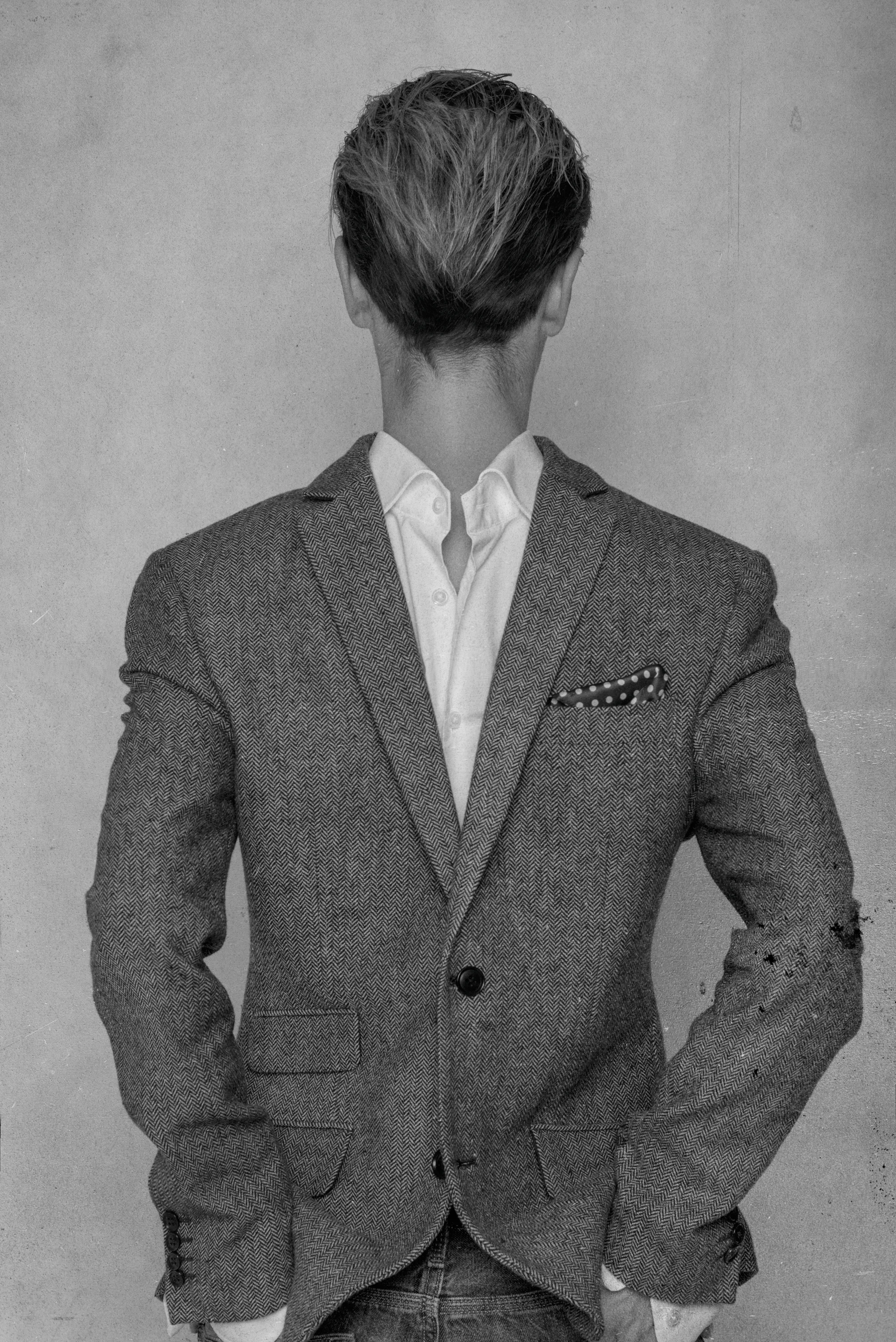 Black-and-white photo of a man wearing a pinstripe blazer over a white shirt, with a polka dot pocket square, standing against a plain wall, facing away from the camera.