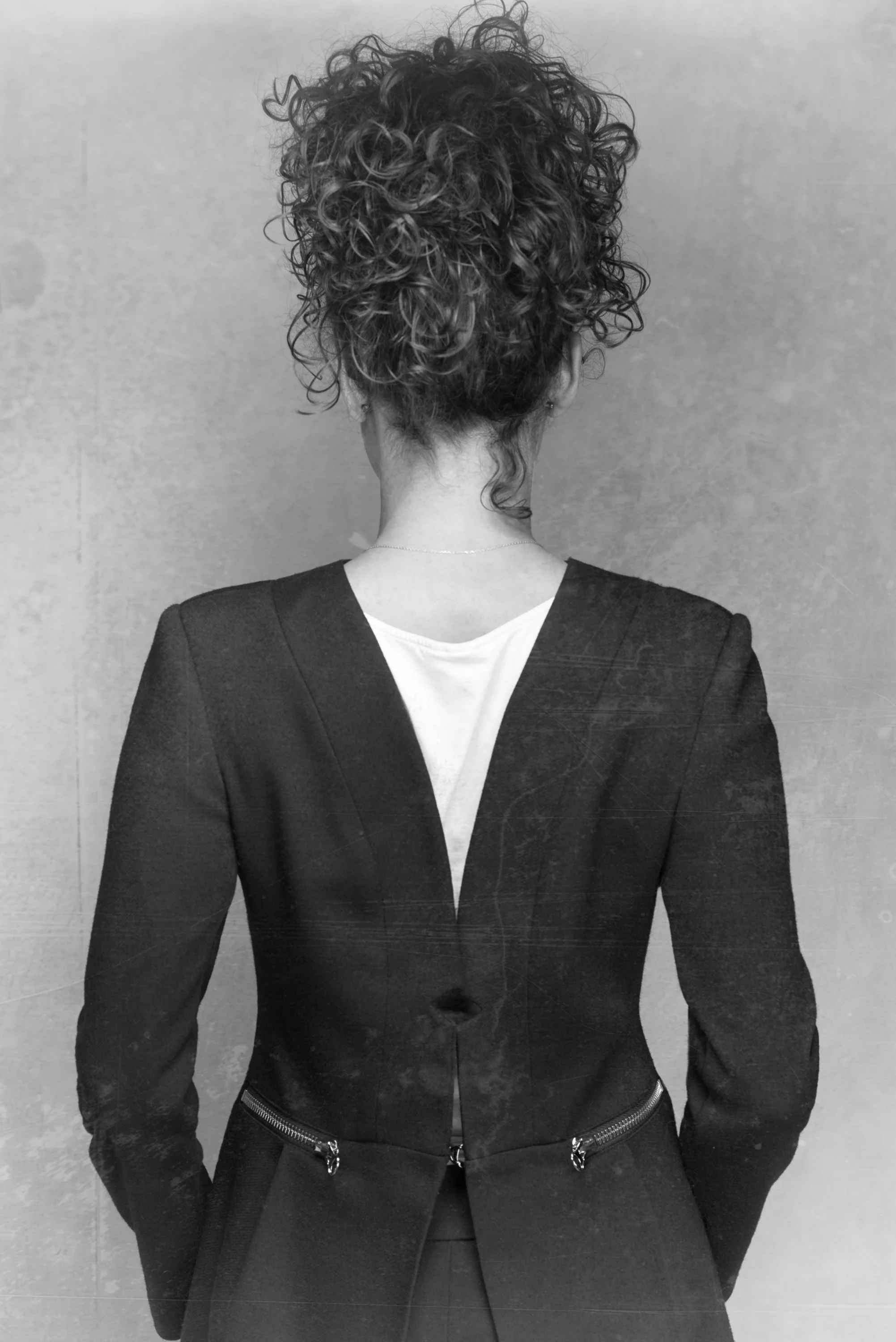 Back view of a woman with short, curly hair, wearing a tailored black blazer with zipper details at the waist, standing against a plain wall.