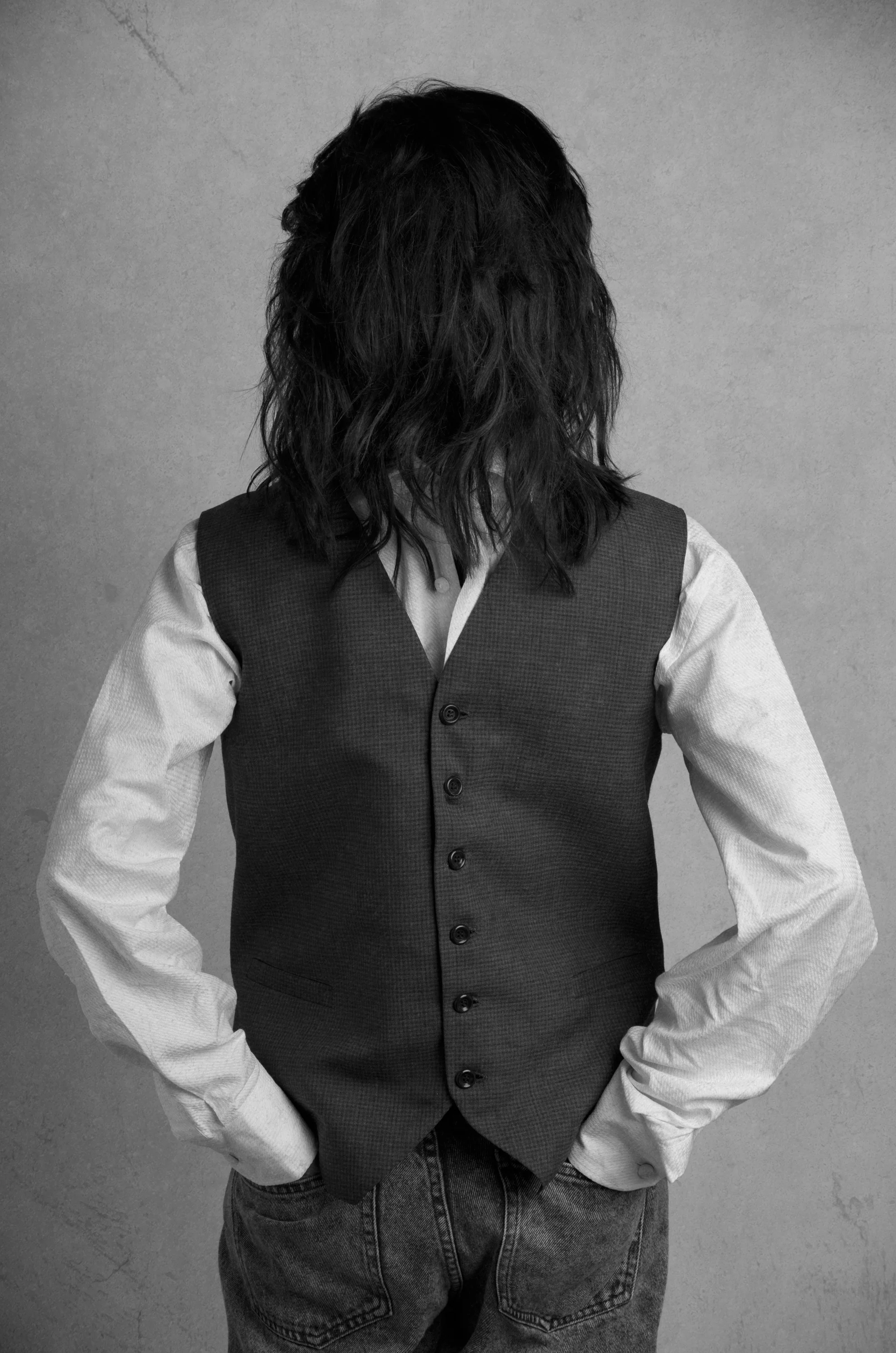 A person with curly dark hair wearing a vest and white shirt, facing away from the camera against a textured gray wall.