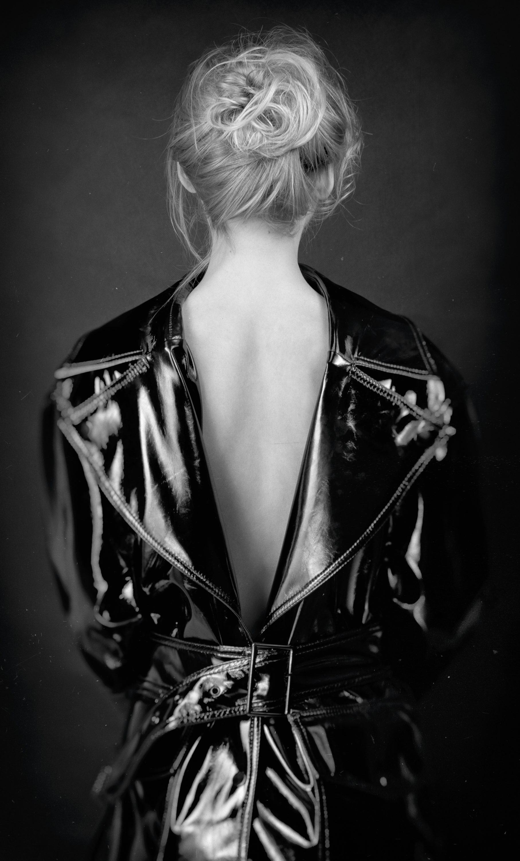 Black and white photograph of a woman with her back facing the camera, her hair styled in an updo, wearing a shiny, black leather jacket with zippers and studs.