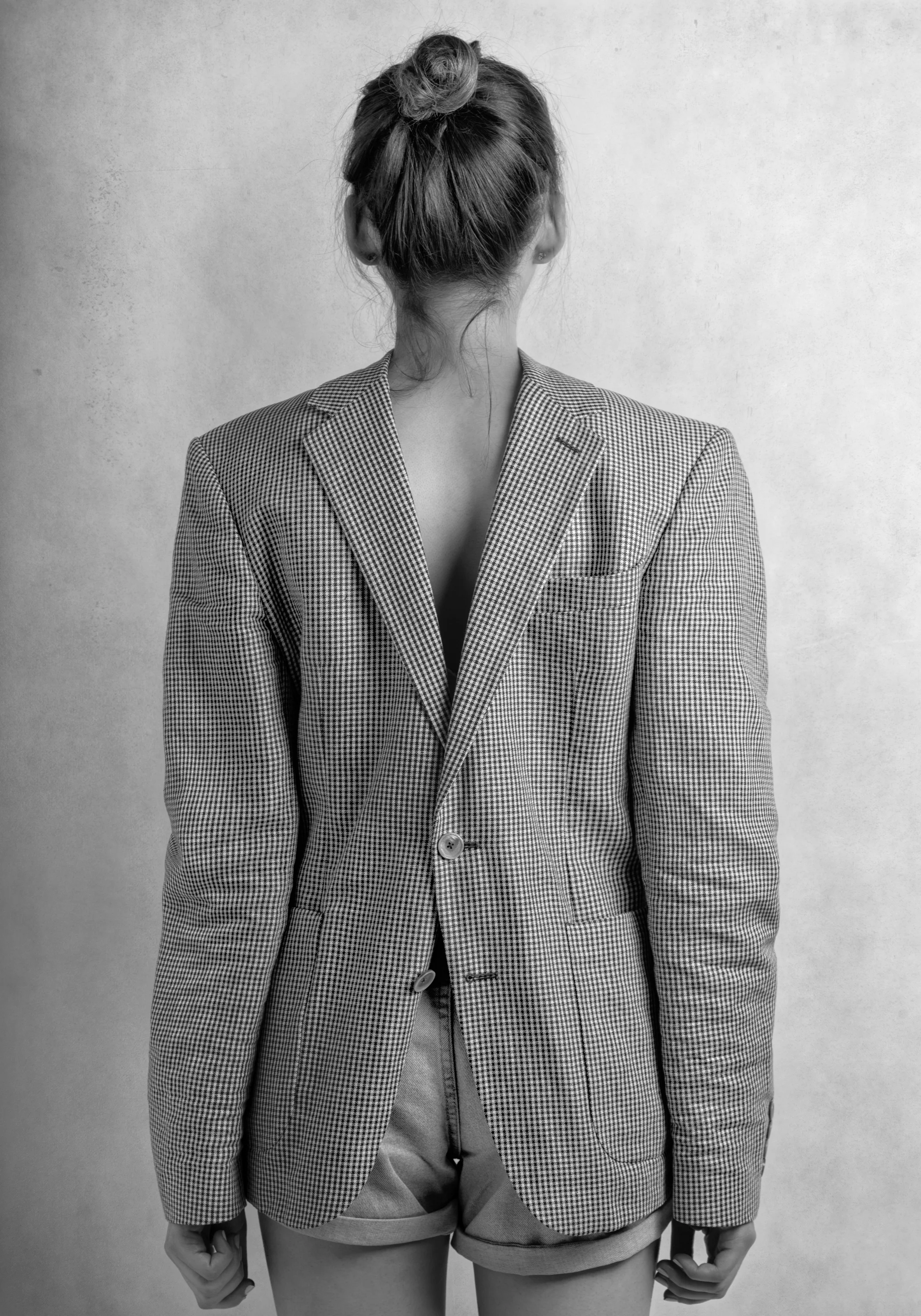A woman with her back to the camera, wearing a checkered blazer and shorts, with her hair in a bun, against a plain wall.