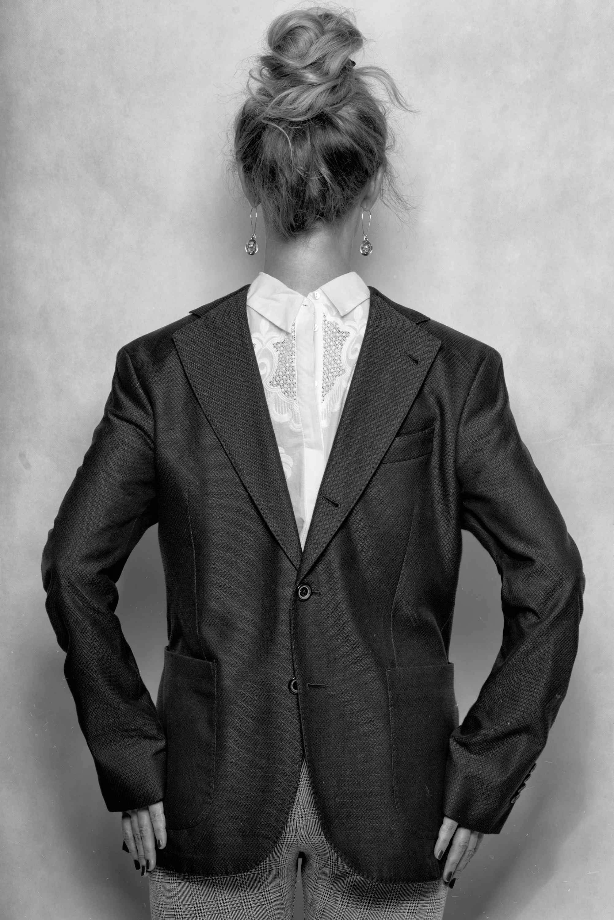 Black and white photo of a woman in a suit with her back to the camera, hair in a messy bun, wearing earrings and plaid pants.