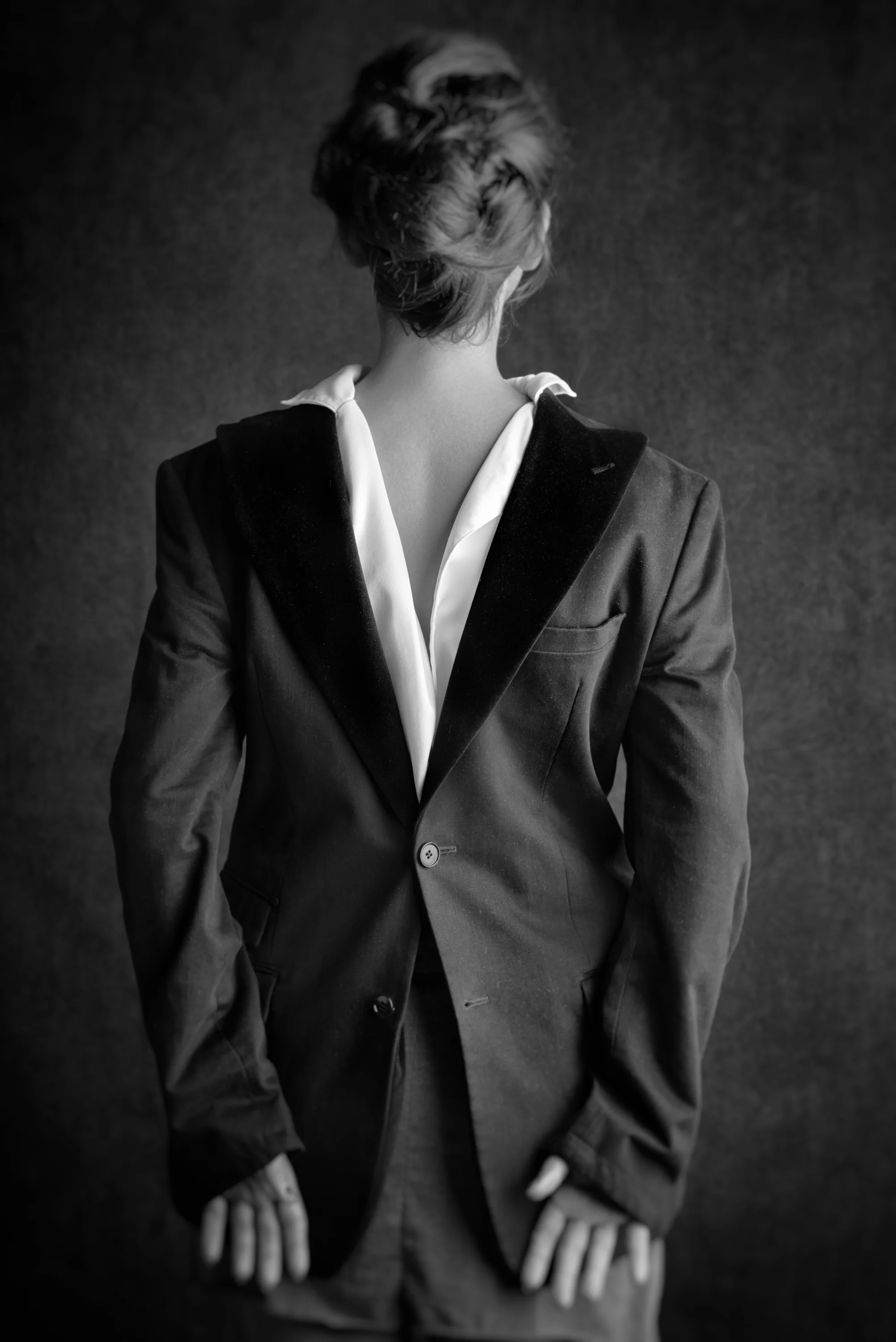 Black and white photo of a woman in a tailored suit, seen from the back, with her hair styled up, against a dark background.