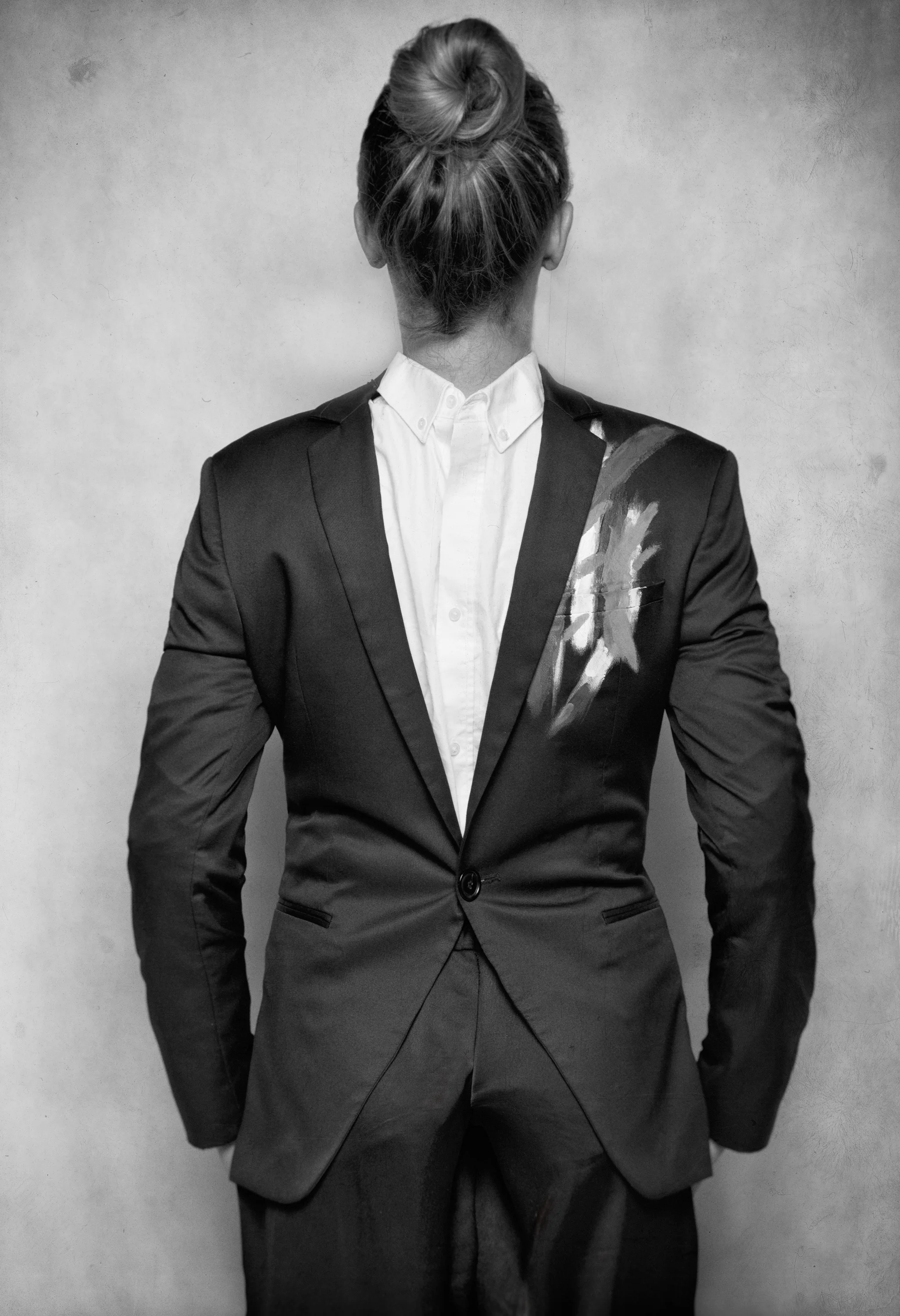 Black and white photo of a person in a suit with a painted blazer, standing against a plain wall. The person's hair is tied up in a bun, and their back is facing the camera.