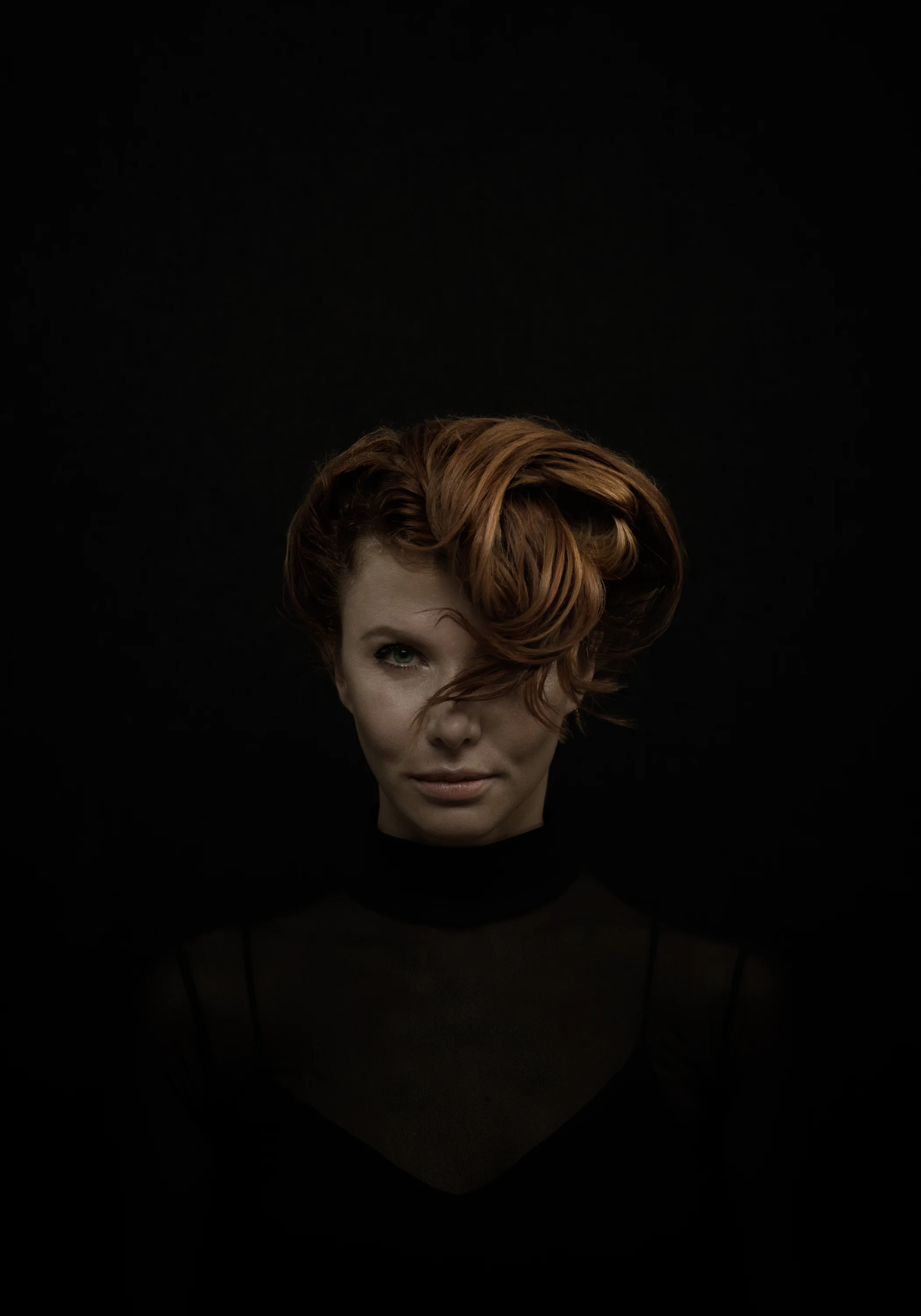 Portrait of a woman with red hair, green eyes, and fair skin, wearing a black sheer top, against a black background.