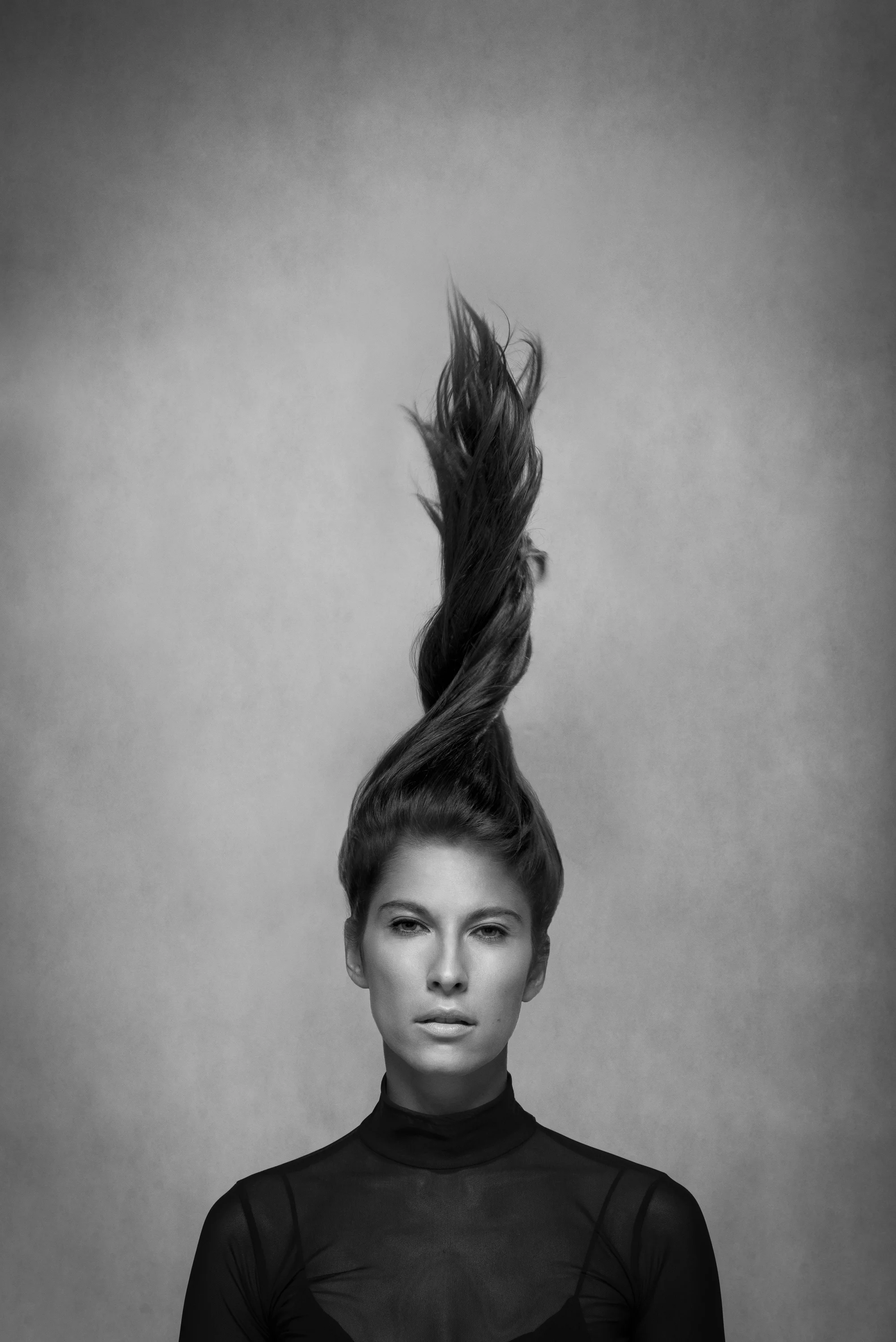 Black and white portrait of a woman with hair lifted upward, wearing a sheer black top, looking directly at the camera.