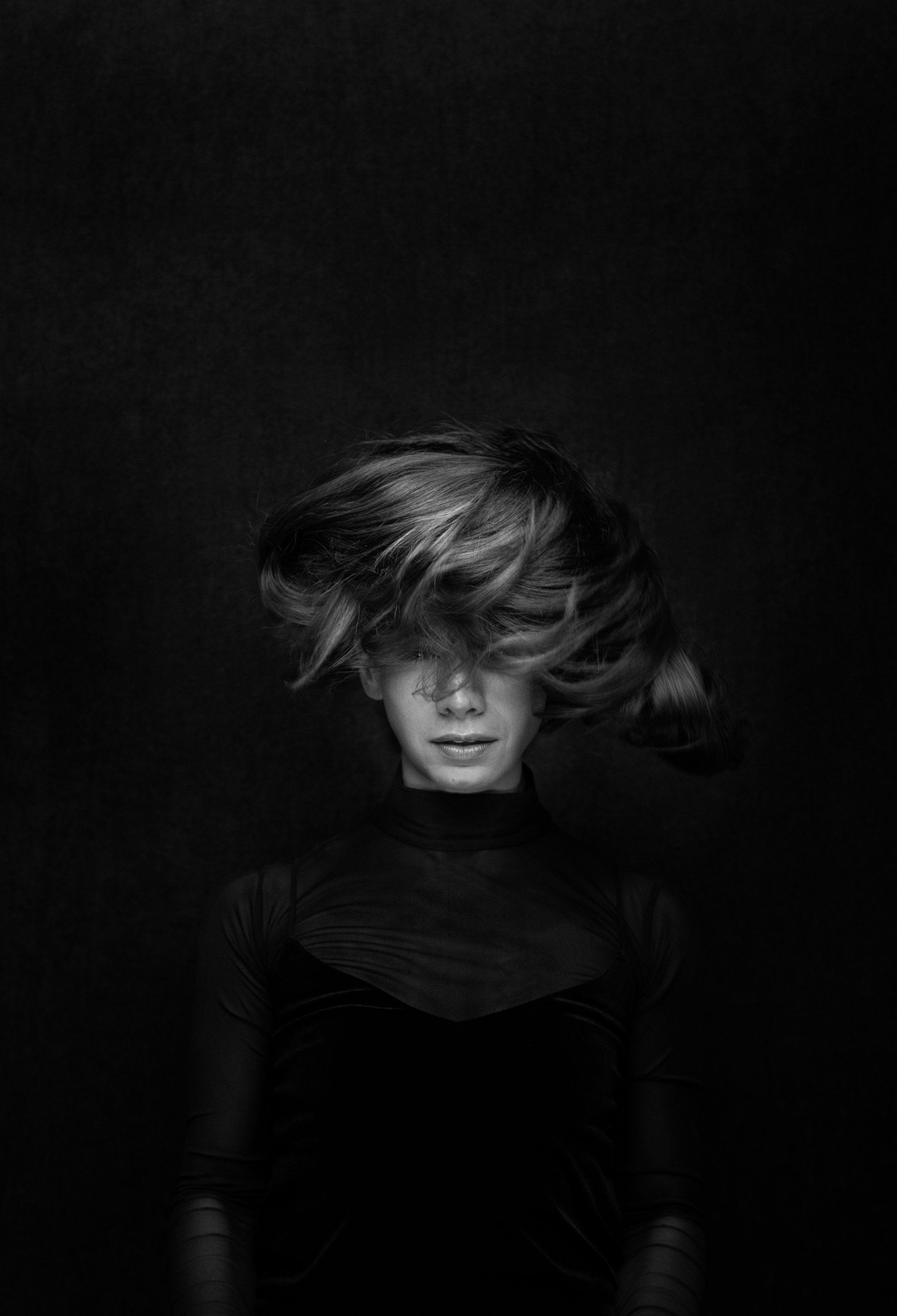 Black and white photo of a woman with short, wavy hair, wearing a fitted, long-sleeve top, looking down with her eyes closed against a dark background.