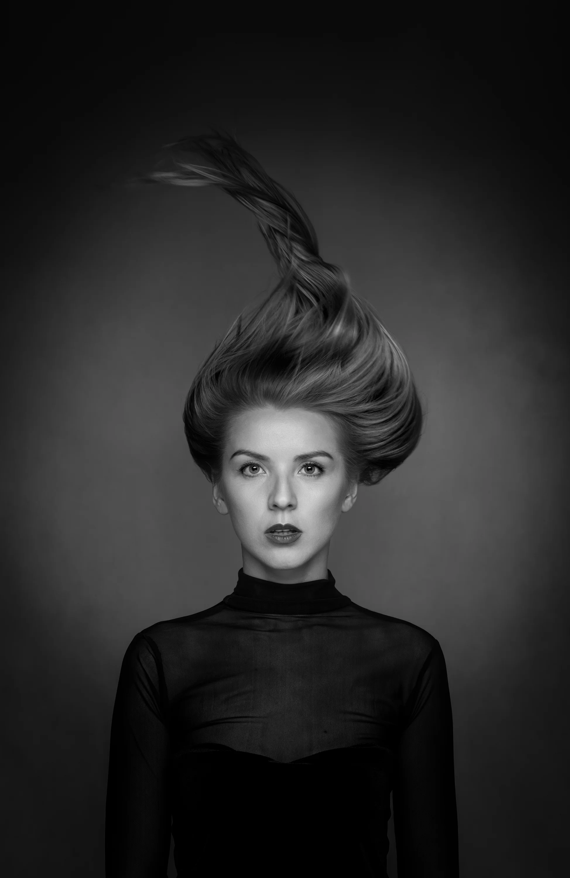 Black and white portrait of a woman with styled hair, wearing a sheer black top, looking directly at the camera against a dark background.