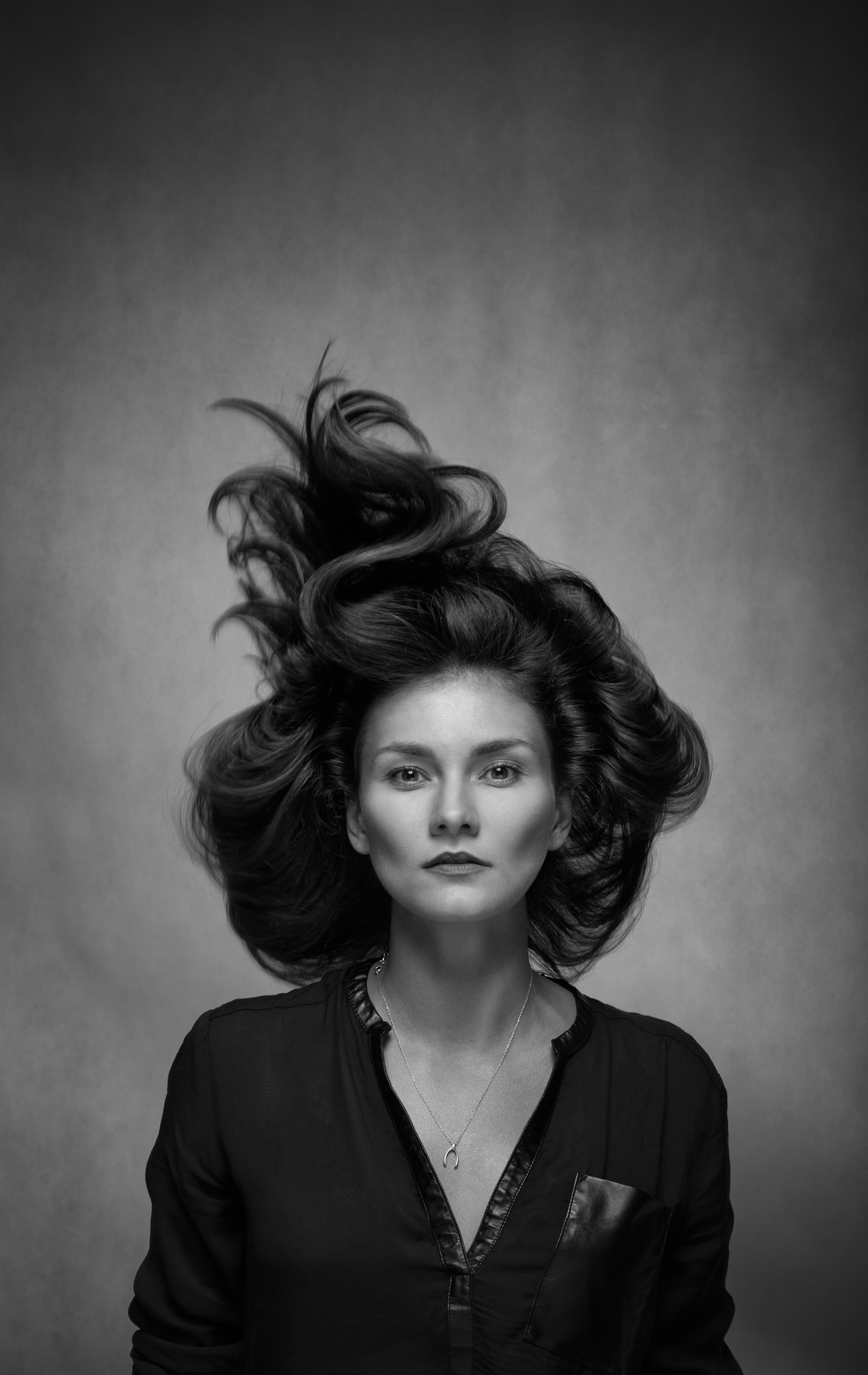 Black and white portrait of a woman with voluminous, wavy hair spread out around her head, wearing a dark top with a zipper and a necklace, against a plain background.