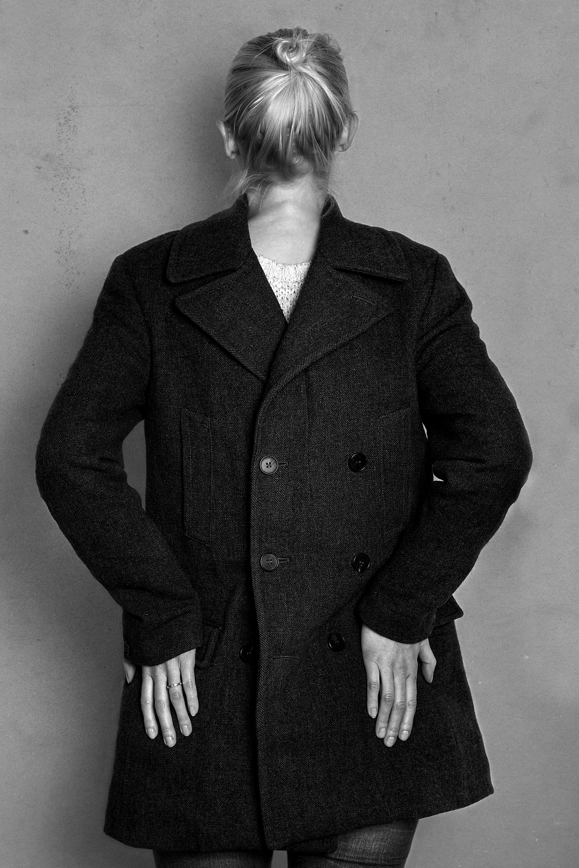 Black and white photo of a woman with her back turned, wearing a dark coat with large buttons and textured fabric, and showing her hands at her sides.