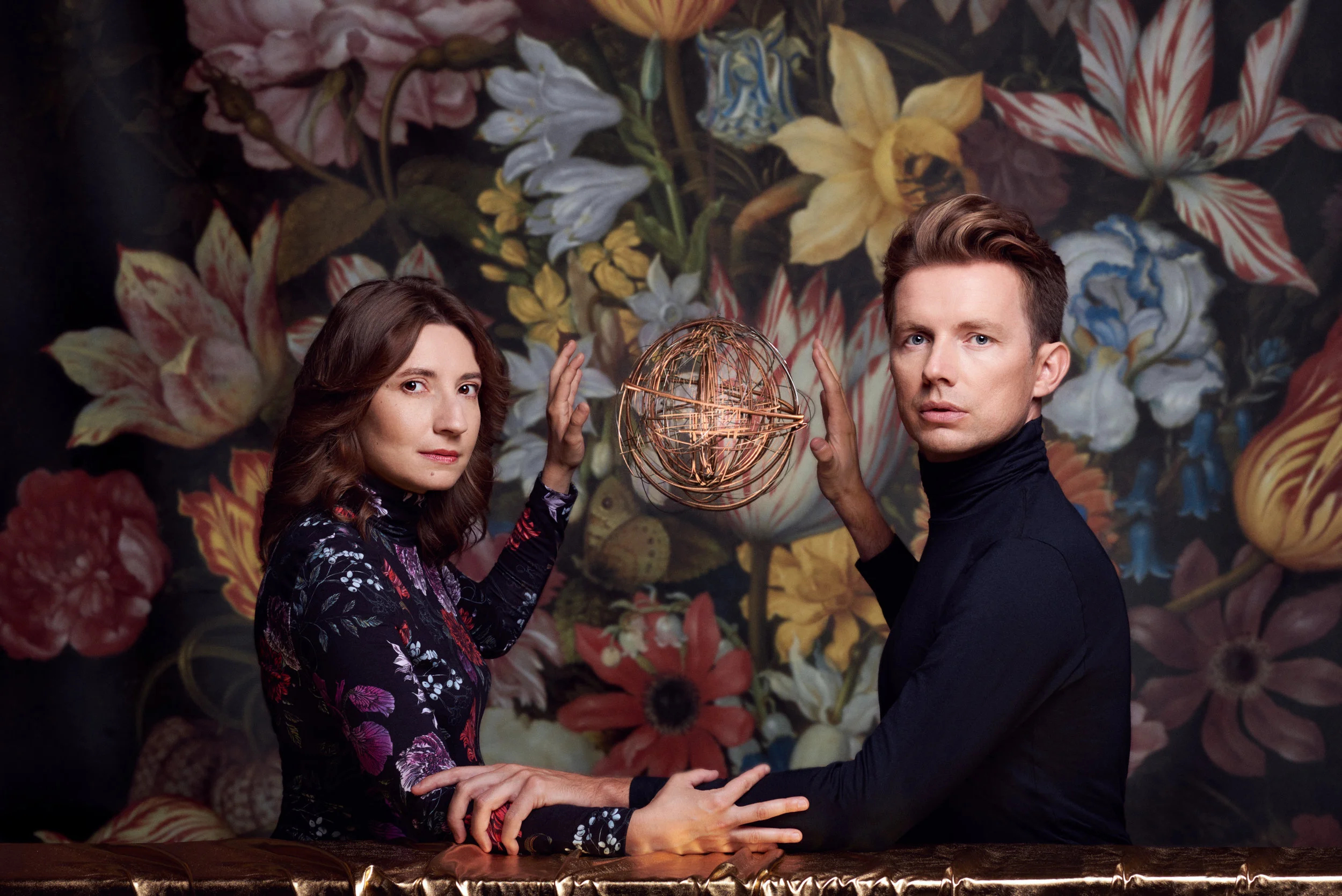 A man and woman are standing close together, holding hands with a decorated wire globe between them, against a floral background. The woman has brown hair and is wearing a dark floral dress, while the man has short brown hair and is wearing a black turtleneck.