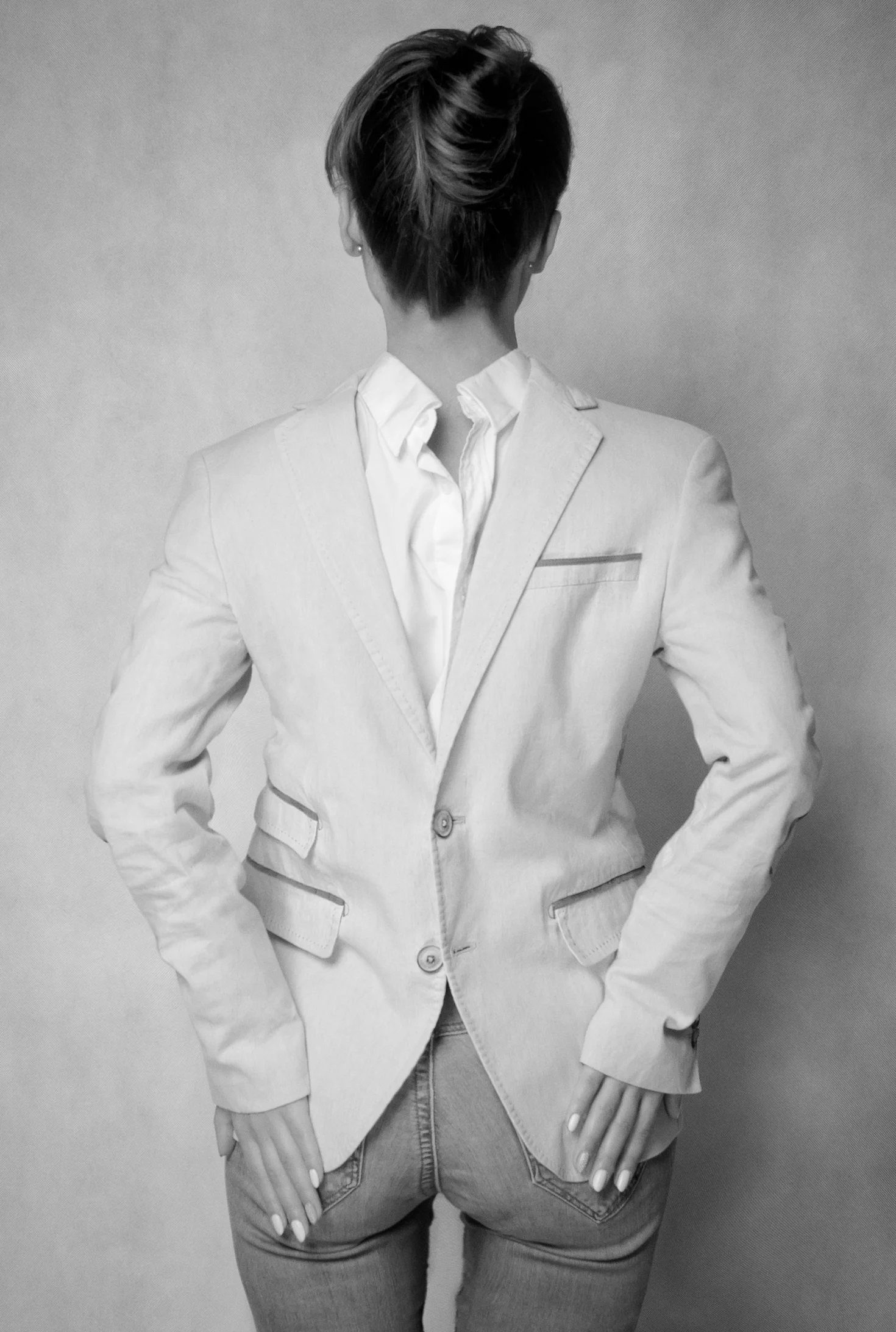 A woman with short hair, wearing a blazer and jeans, is standing with her back to the camera against a plain wall.