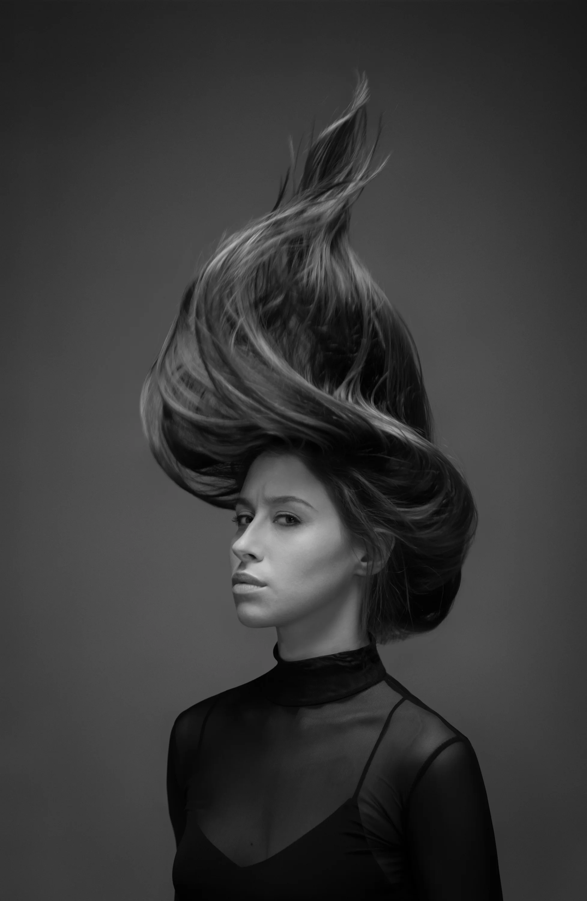 A black and white portrait of a woman with hair flying upward and to the side, wearing a high-neck black top with sheer fabric on the shoulders, against a dark background.