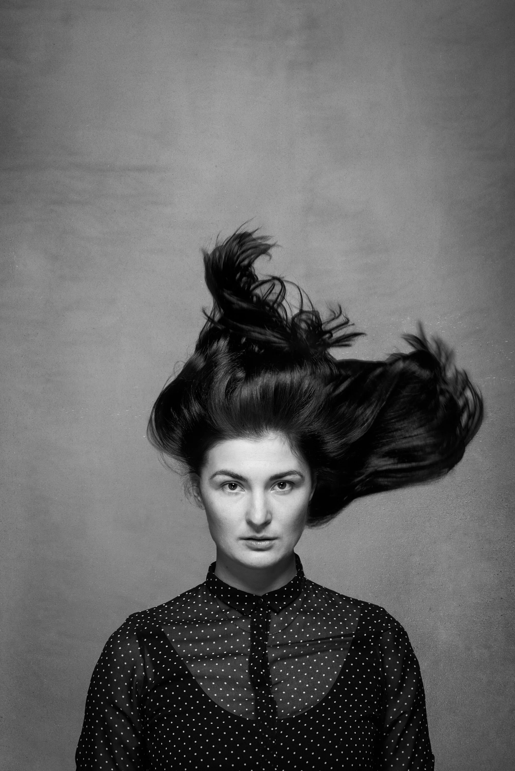 A black and white portrait of a woman with long, flowing dark hair, wearing a polka dot shirt, against a plain background.