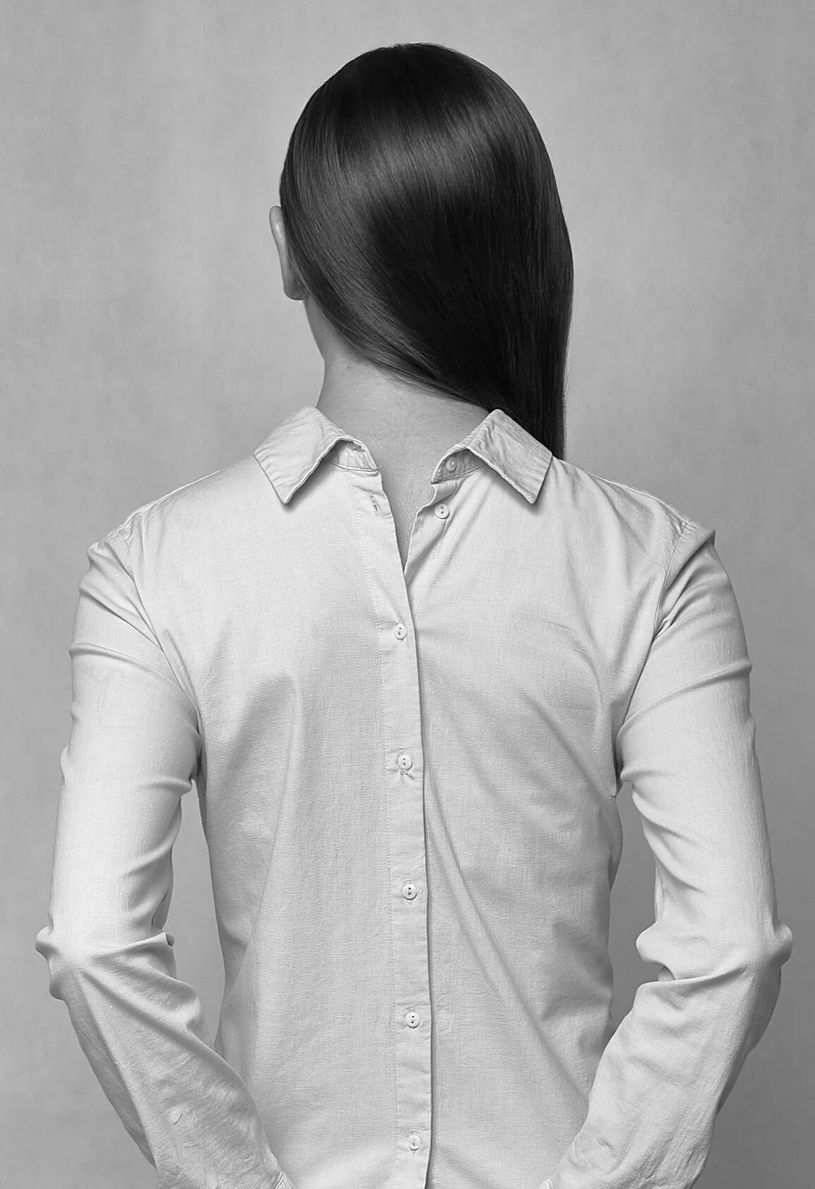 Black and white photo of a woman with straight, shoulder-length hair, facing away, wearing a light-colored button-up shirt with rolled-up sleeves.