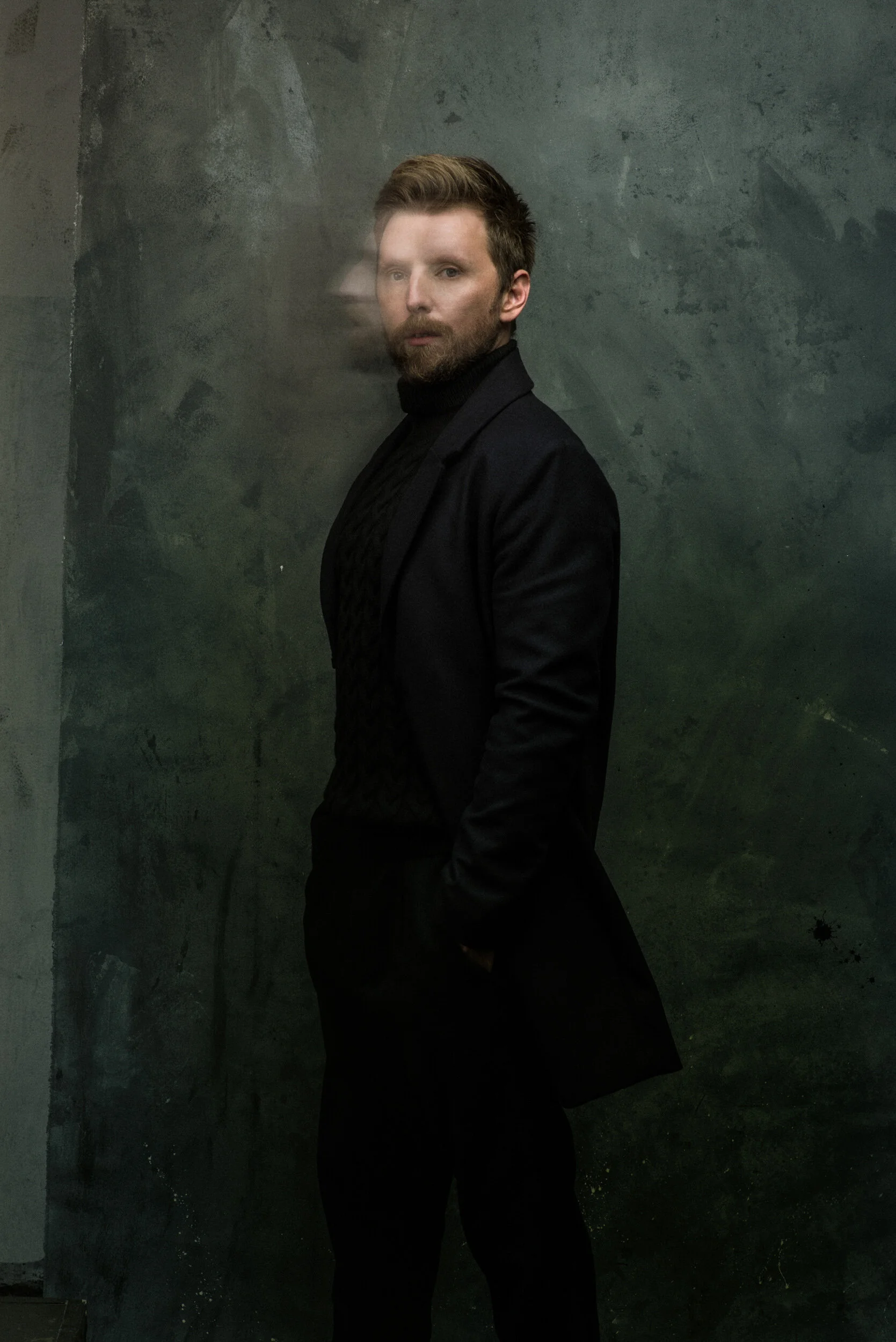A man with brown hair and a beard wearing a black turtleneck and black blazer, standing sideways against a textured dark green wall.