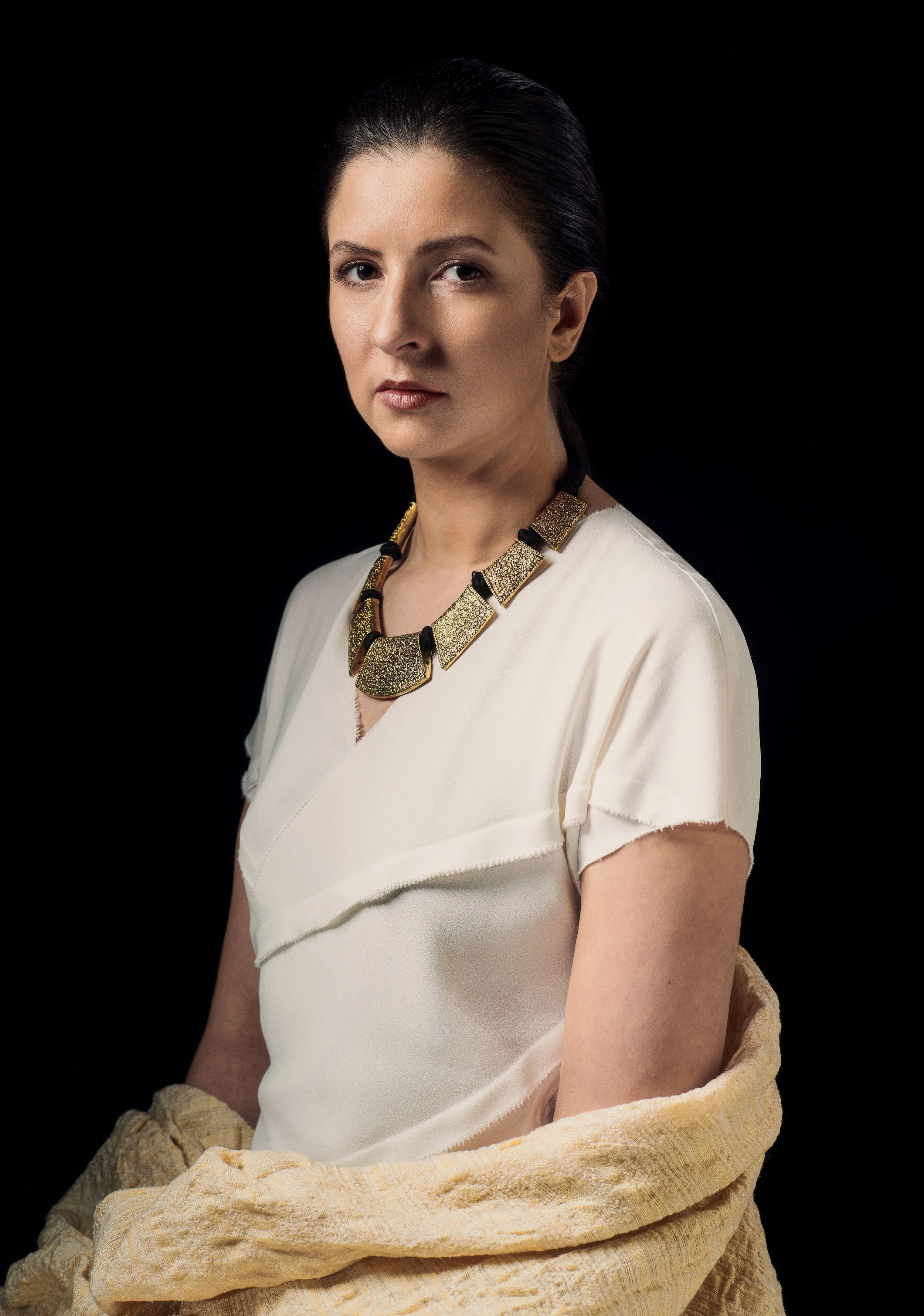A woman with dark hair pulled back, wearing a white dress and a large, textured gold and black necklace, against a solid black background.