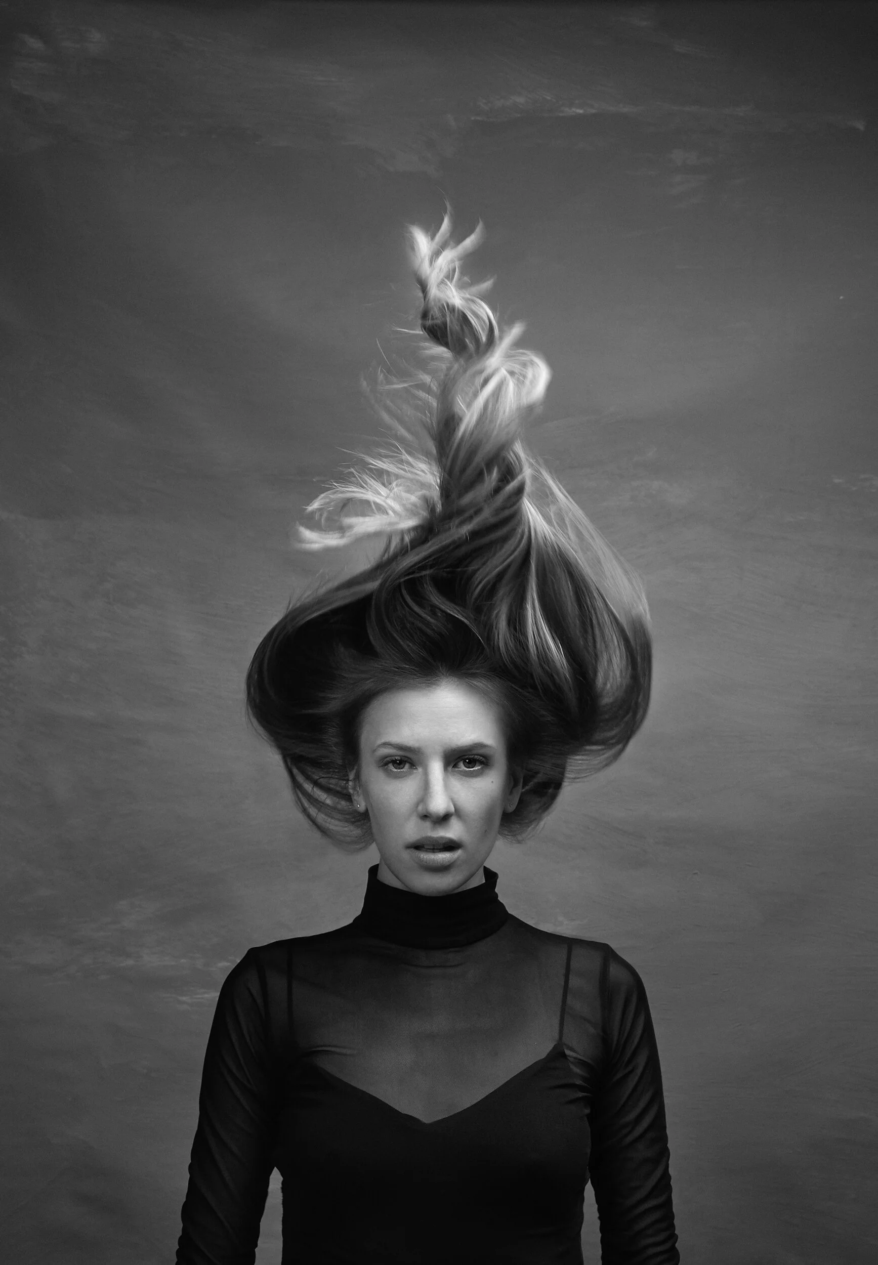 A woman with long hair flying upwards into the air against a cloudy sky, wearing a black, sheer long-sleeve top and looking directly at the camera.