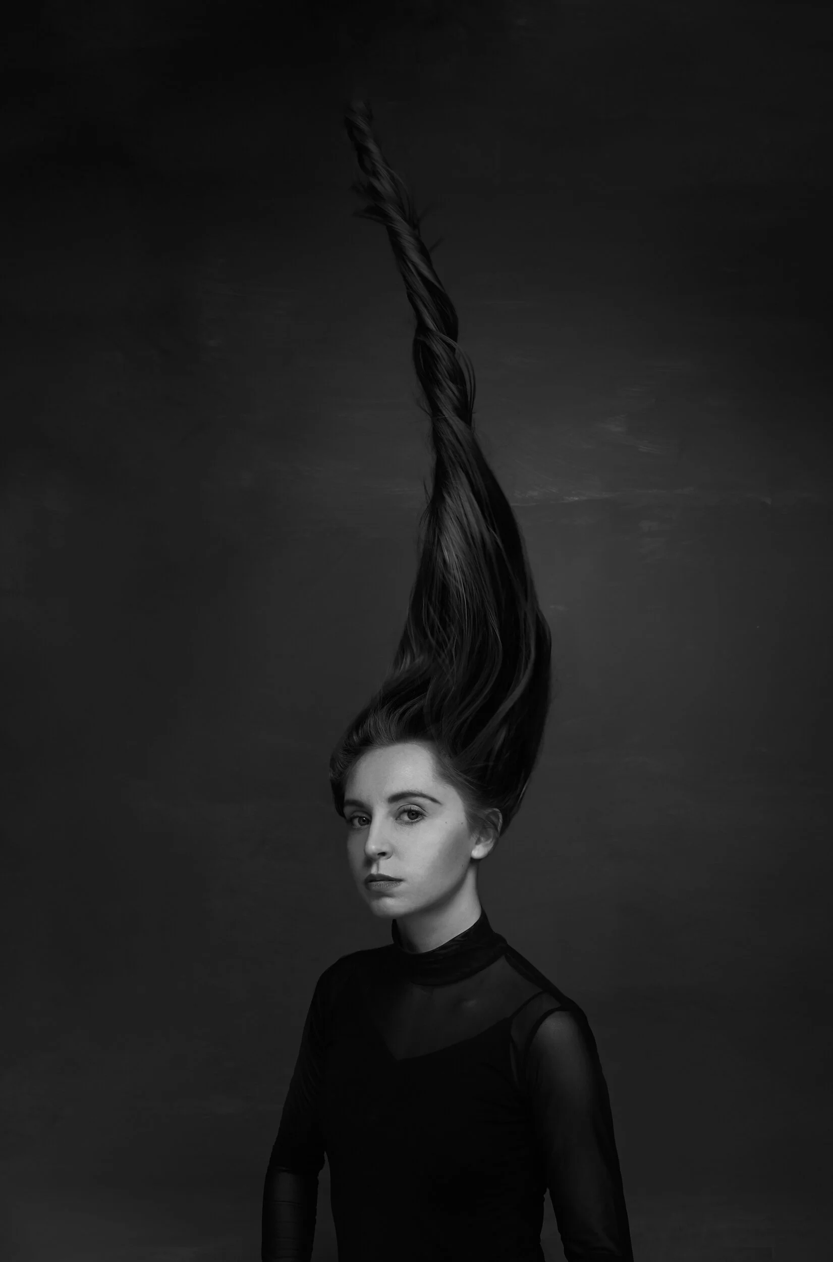 Black and white portrait of a woman with arched eyebrows, wearing a high-neck sheer black top, standing against a dark background, with her face turned slightly to the side and her hair flowing upward.