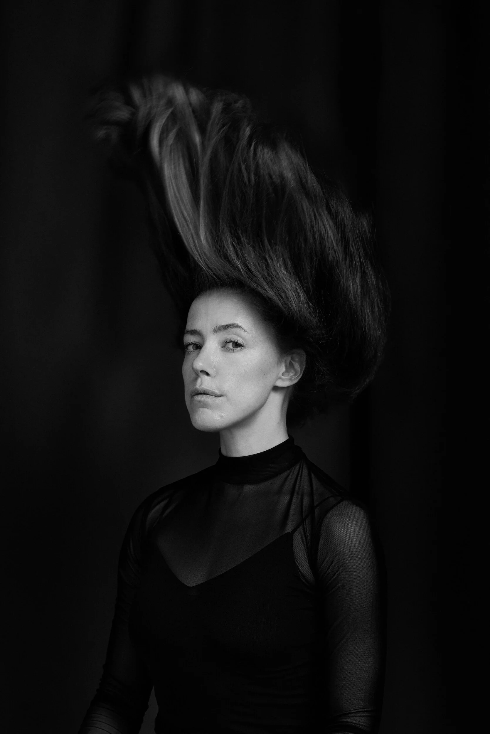 A black and white photograph of a woman with short hair, wearing a dark, sheer, long-sleeved top. Her head is tilted slightly to one side, and her hair is floating upward, creating a dramatic effect against the dark background.