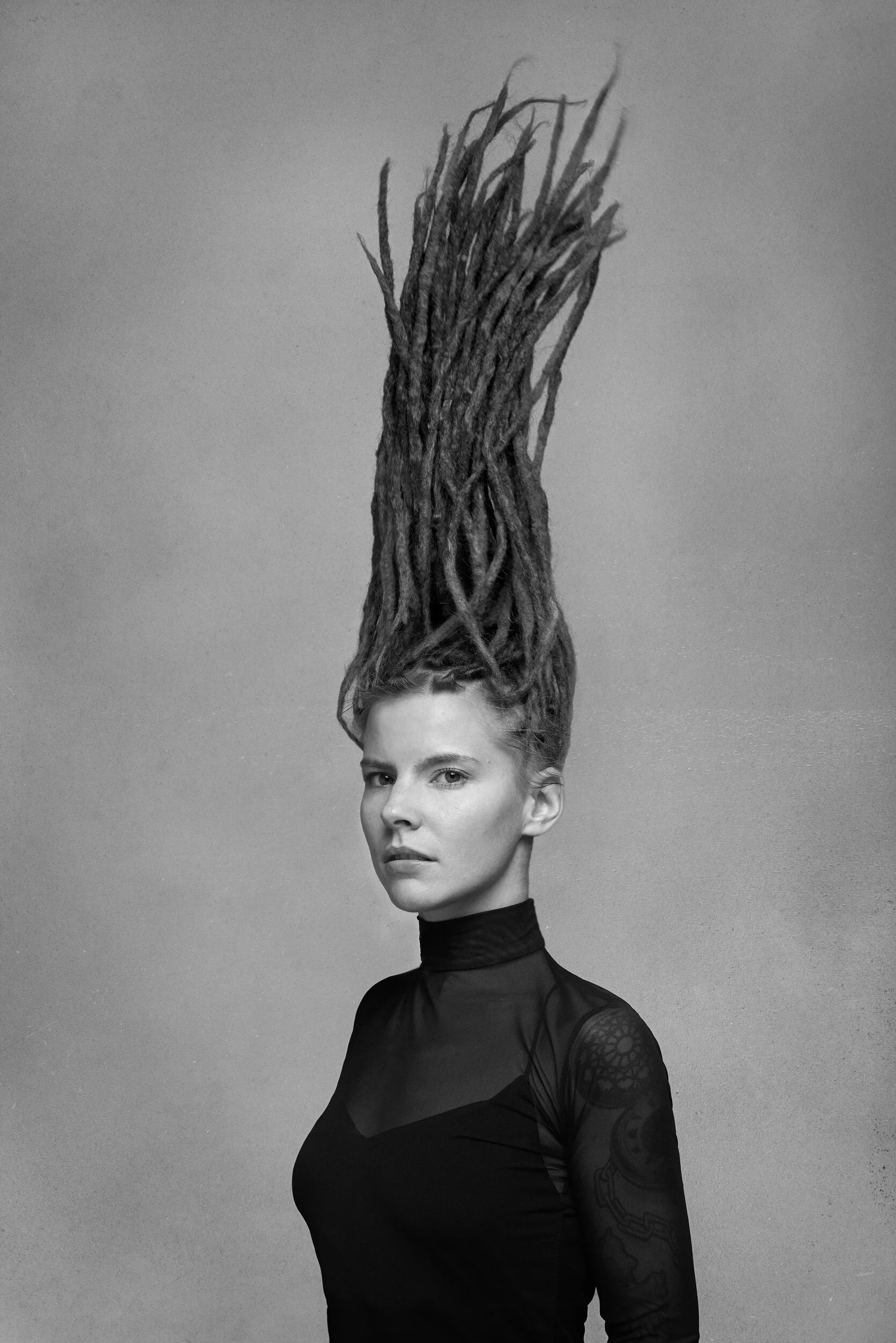 A black and white photo of a woman with a tall hairstyle resembling dreadlocks or hair extensions, wearing a high-neck, long-sleeved lace top, against a plain background.