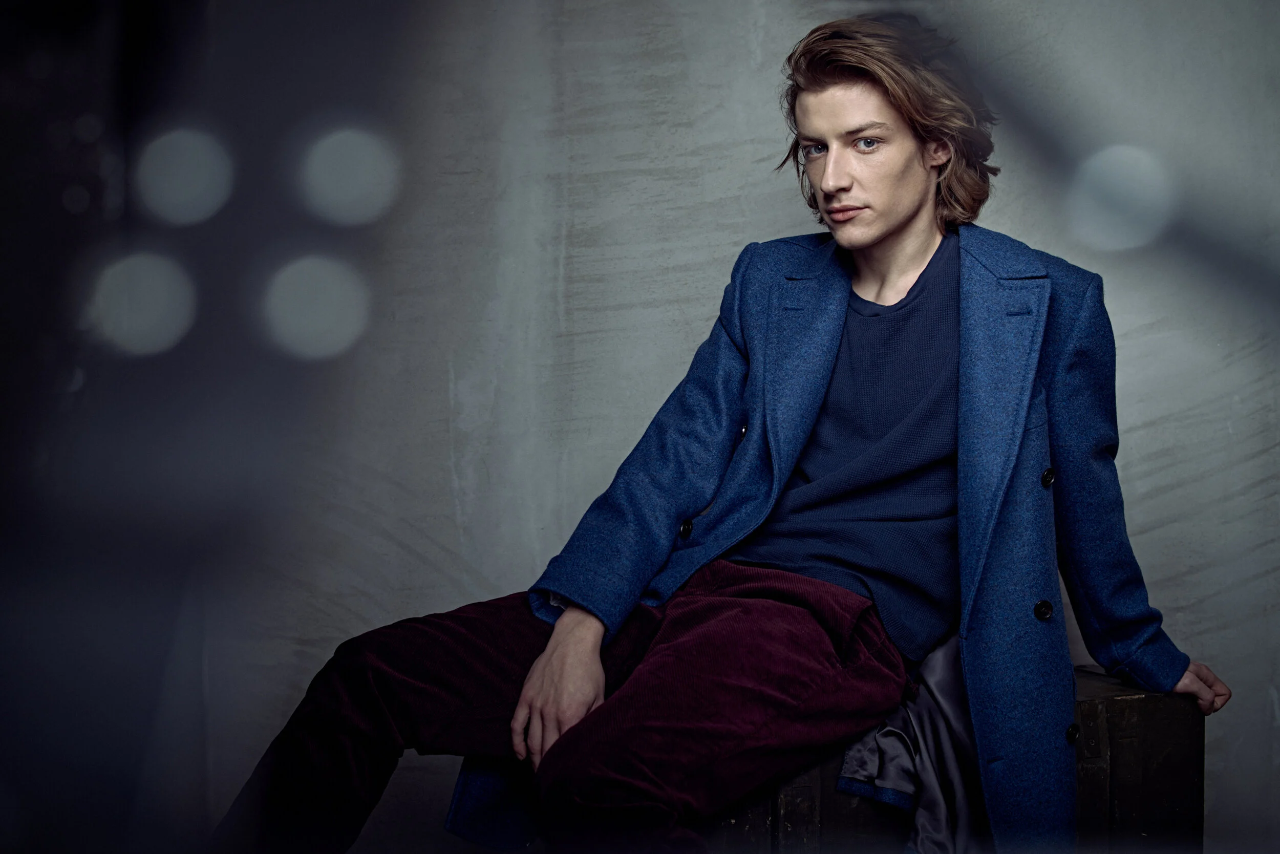A young man with wavy, shoulder-length brown hair, wearing a blue blazer and dark shirt, sitting against a gray wall with bokeh lights in the background.