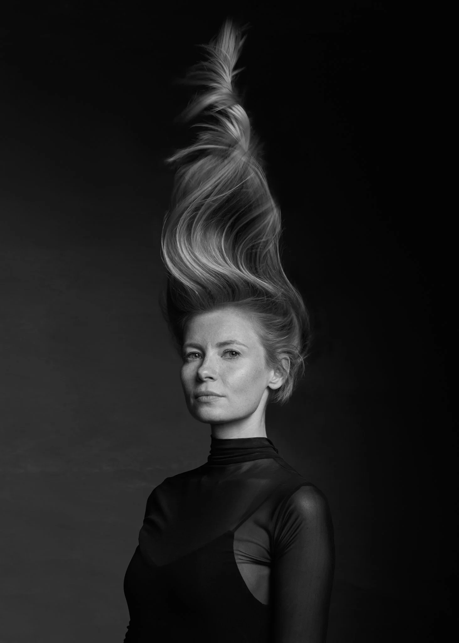 A black-and-white portrait of a woman with short hair wearing a dark, sheer top. Her hair is styled upward and appears to be in motion, with a swirling shape extending above her head. She looks confidently at the camera against a dark background.