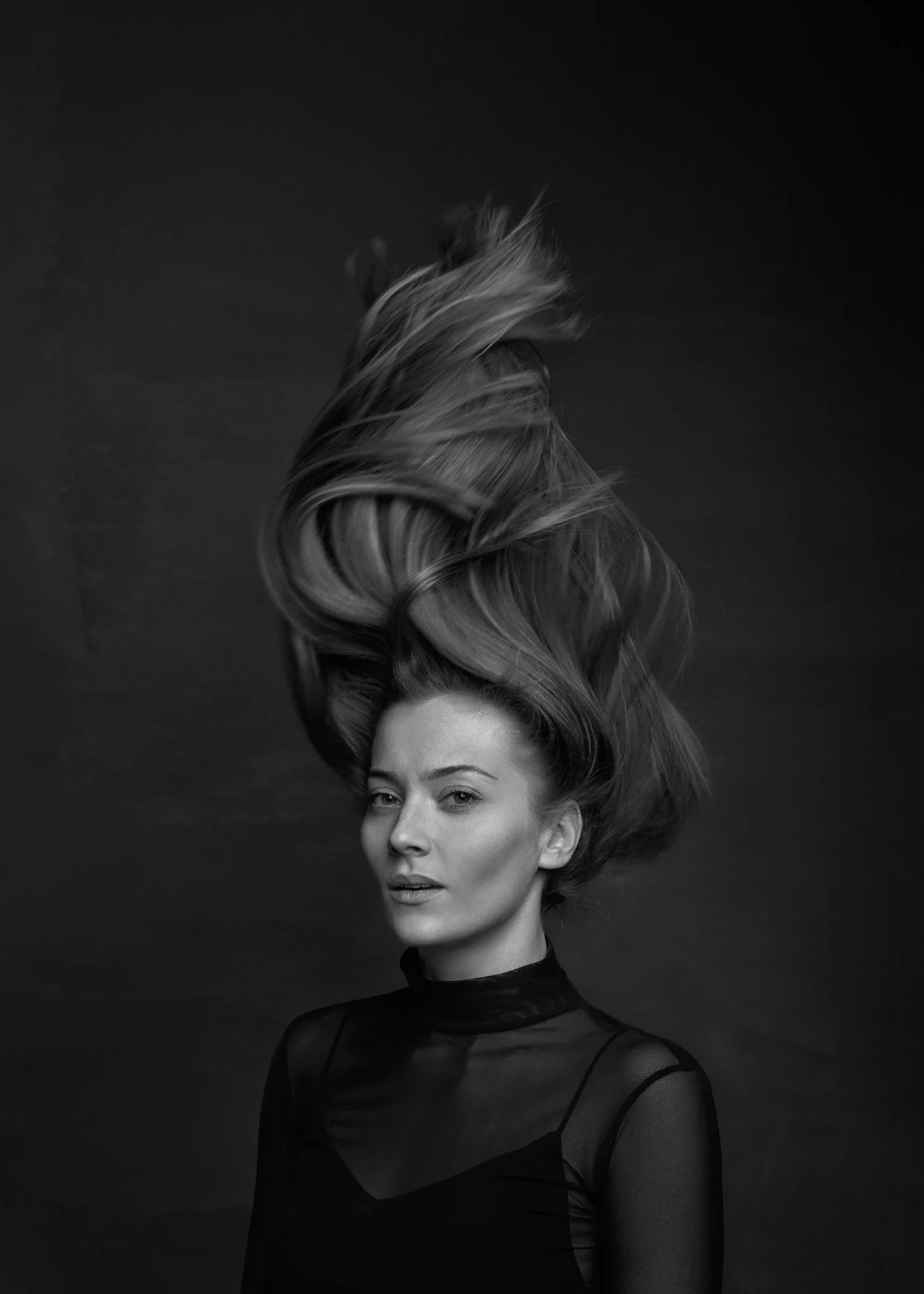 Black and white portrait of a woman with flowing blonde hair, wearing a sheer black top, against a dark background.