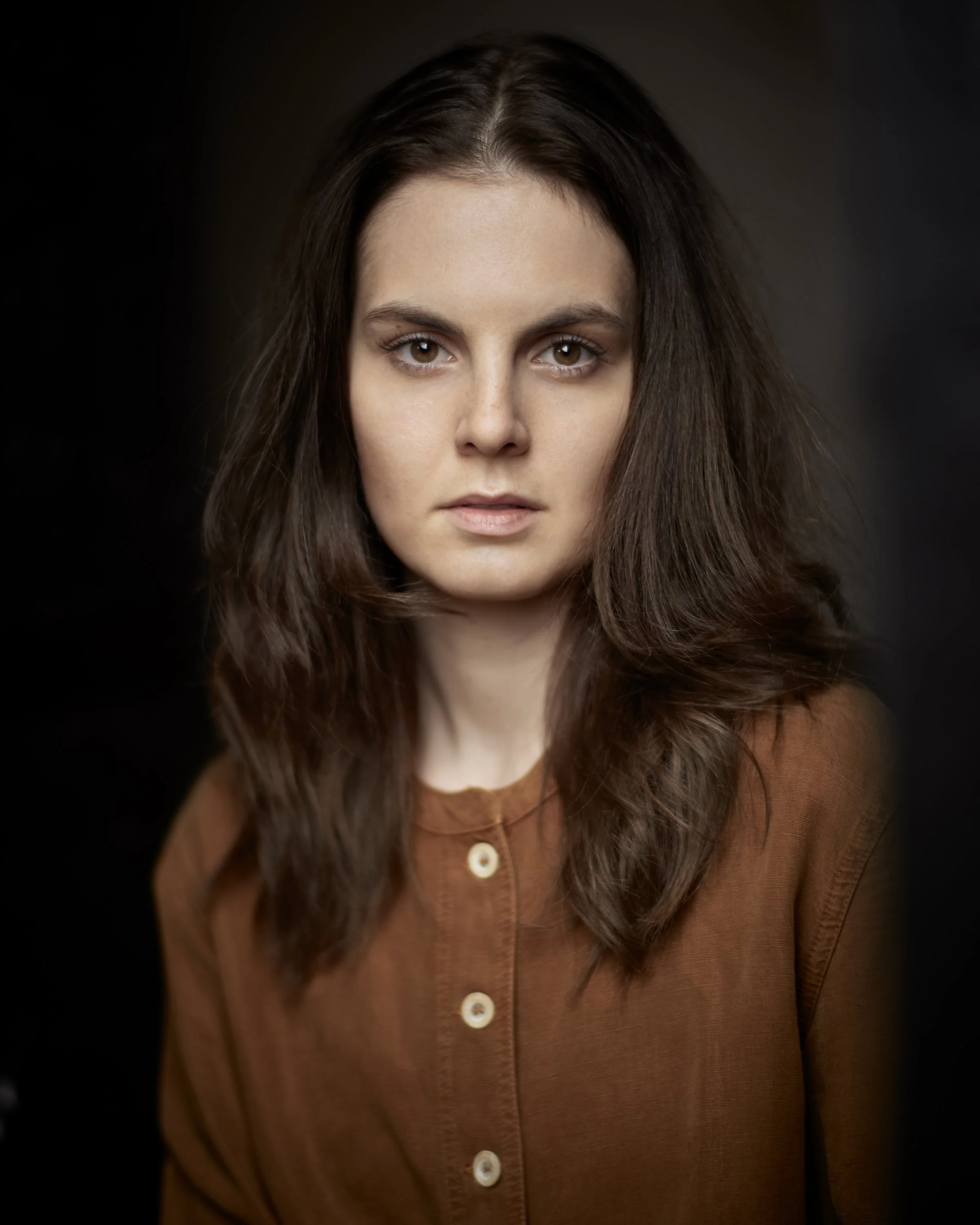 Portrait of a woman with brown hair and brown eyes wearing a brown shirt with buttons, looking directly at the camera against a dark background.