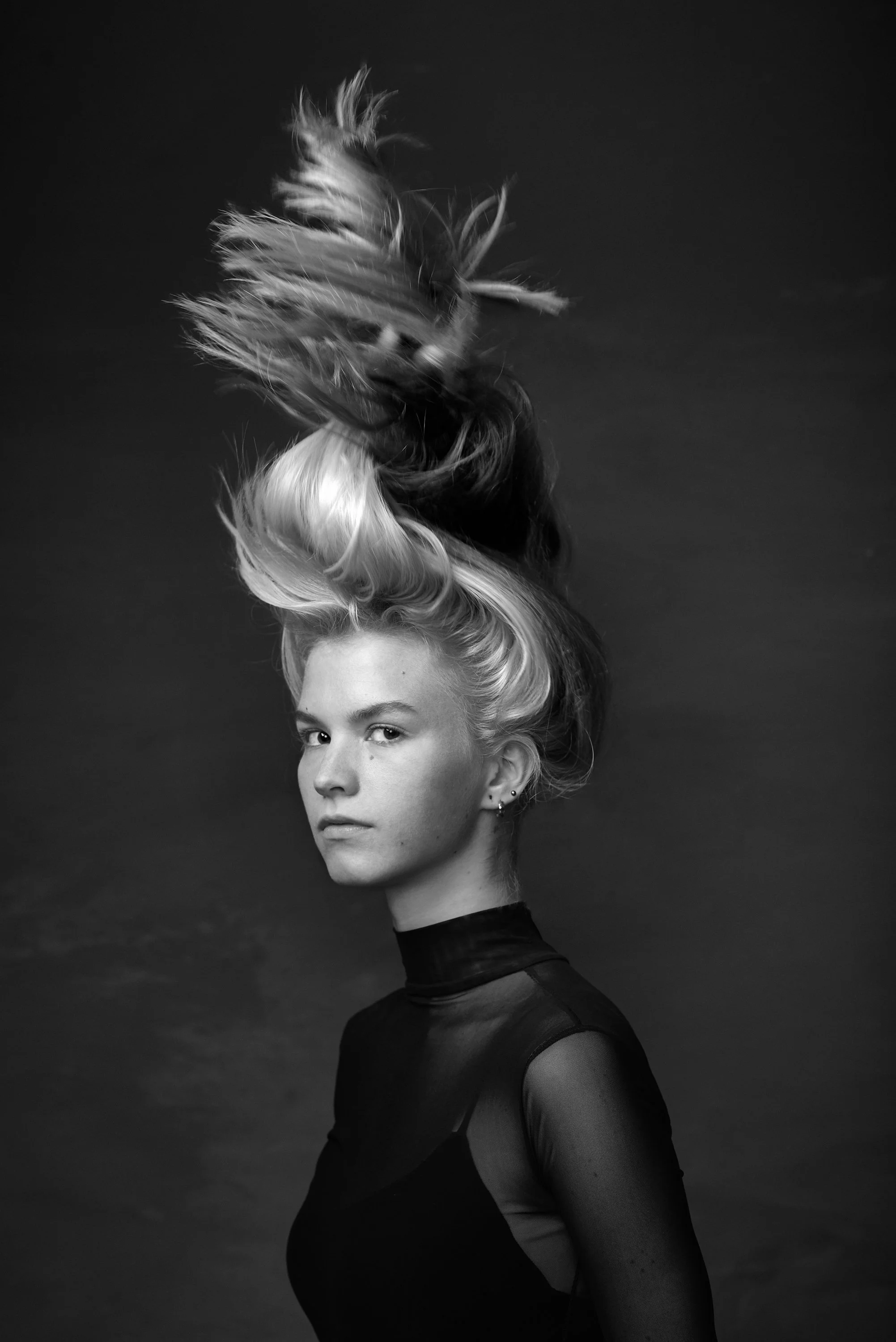 A black and white portrait of a woman with light-colored hair styled in an elaborate, voluminous updo, wearing a high-neck, form-fitting sheer dress with opaque sections.