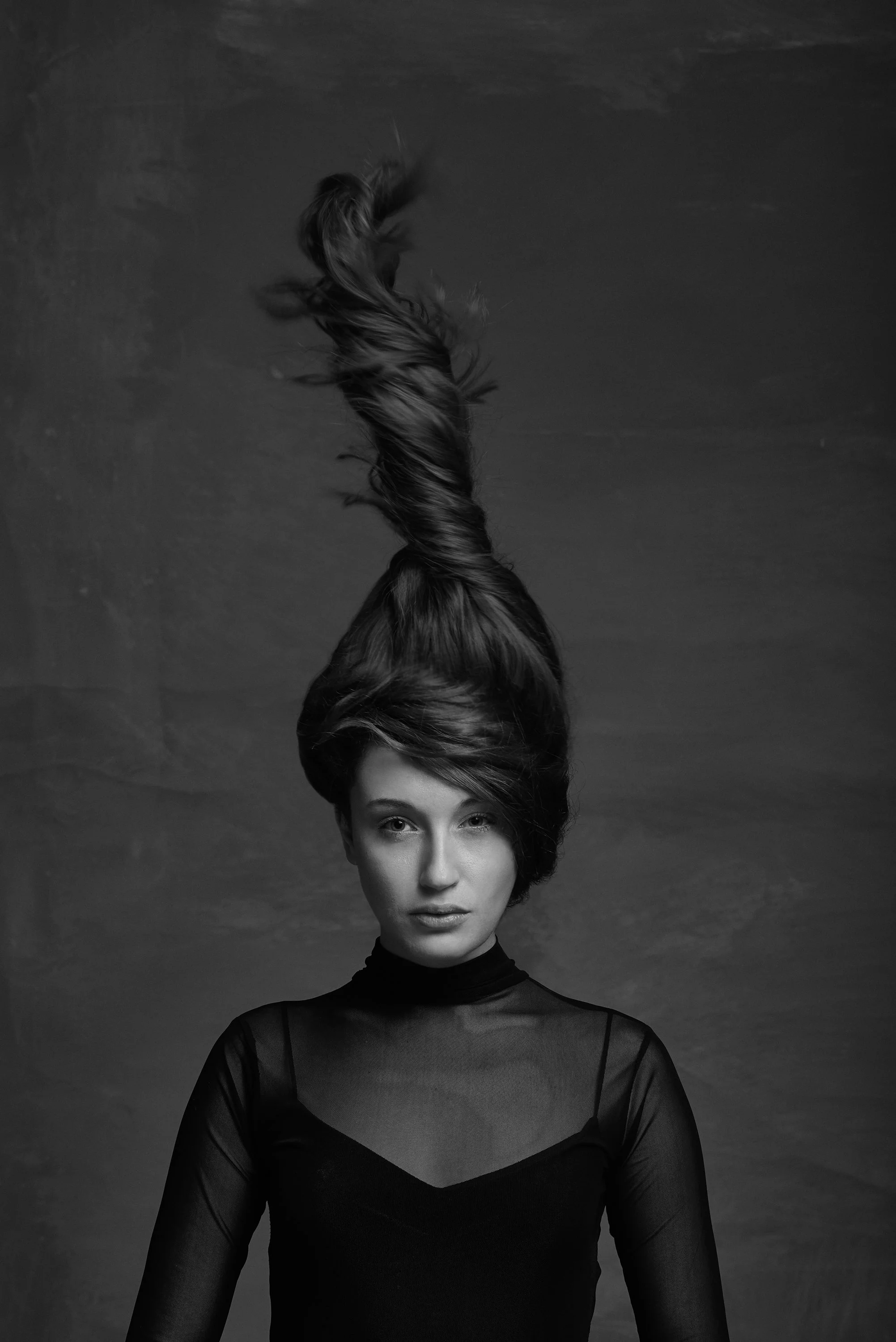 Black and white portrait of a woman with short hair. Her hair is styled in an unusual upward twist with loose strands. She is wearing a sheer black top with a high collar and long sleeves, standing against a textured background.