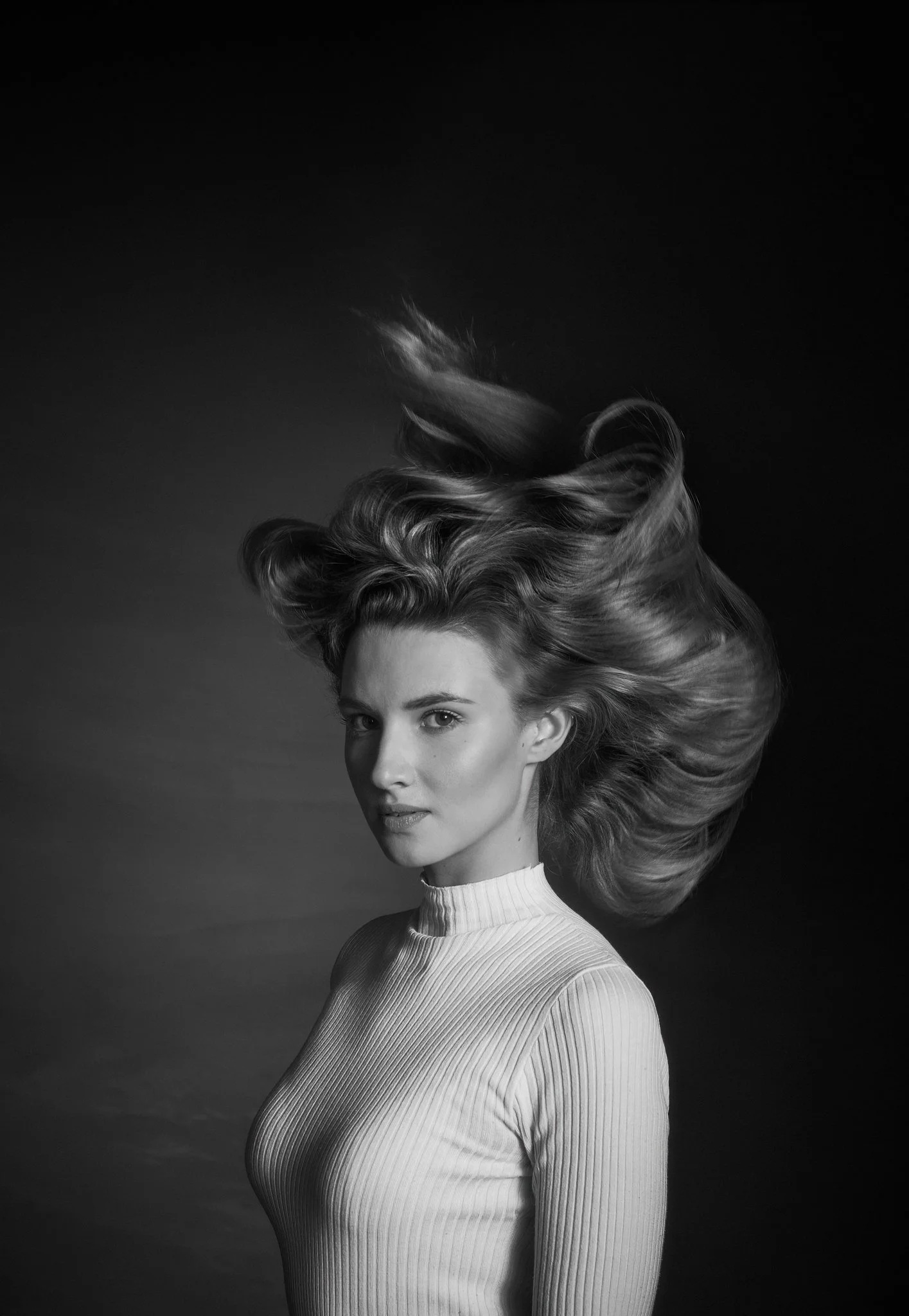 Black and white portrait of a woman with her hair flowing, wearing a ribbed turtleneck sweater, looking at the camera.