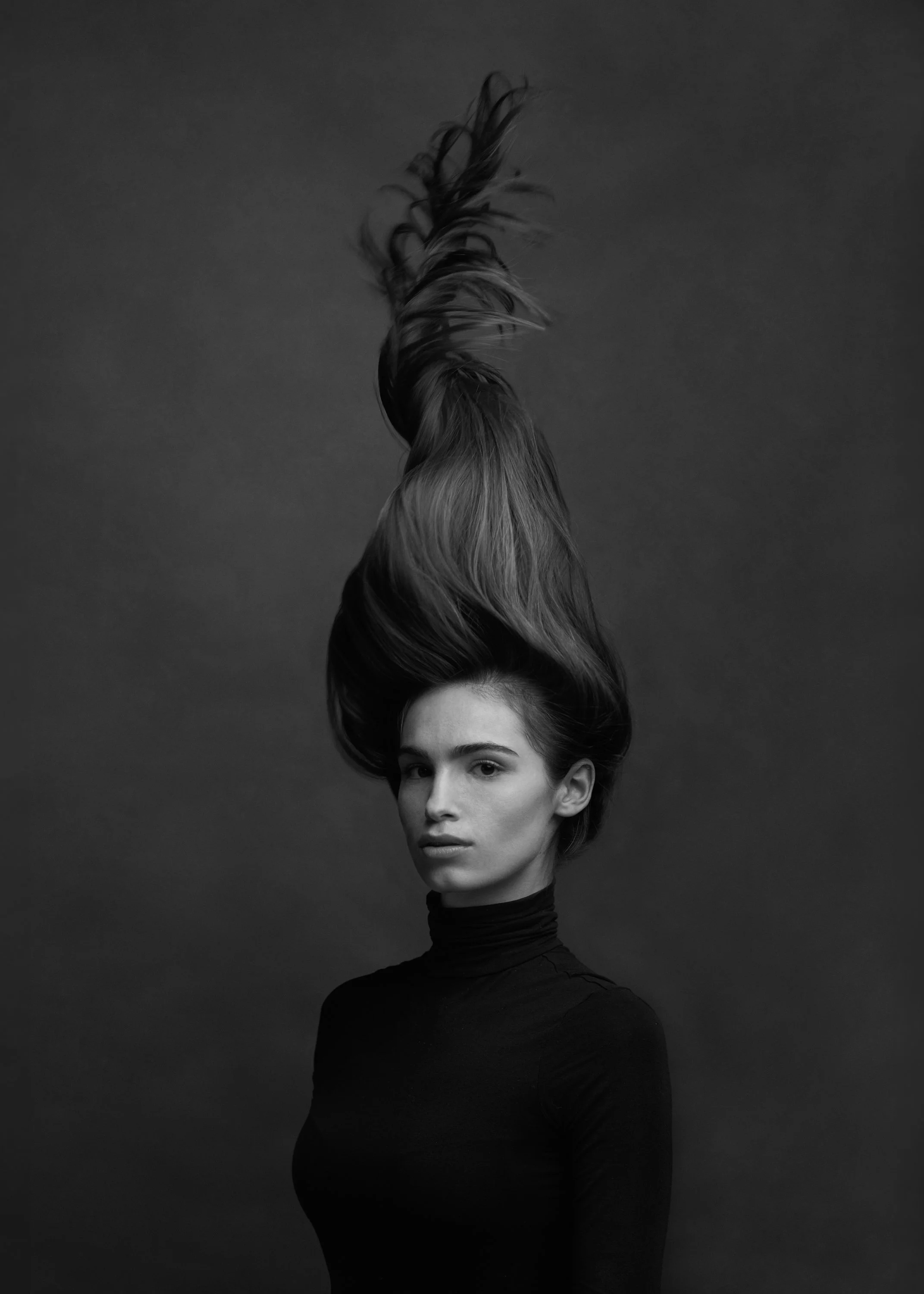 Black and white portrait of a woman with long hair styled upward, wearing a black turtleneck, against a plain dark background.