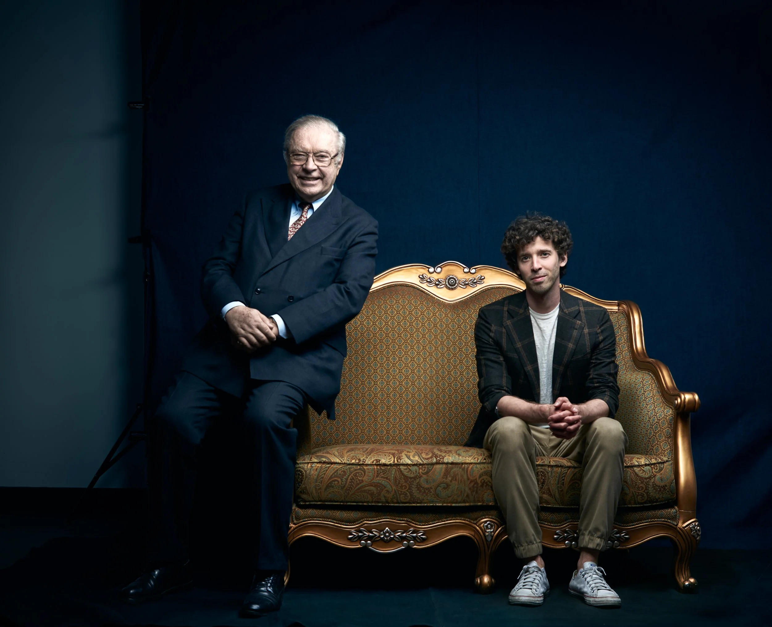 Older man in a business suit shaking hands with a younger man in casual clothes, sitting on an ornate gold and fabric sofa against a dark blue wall.