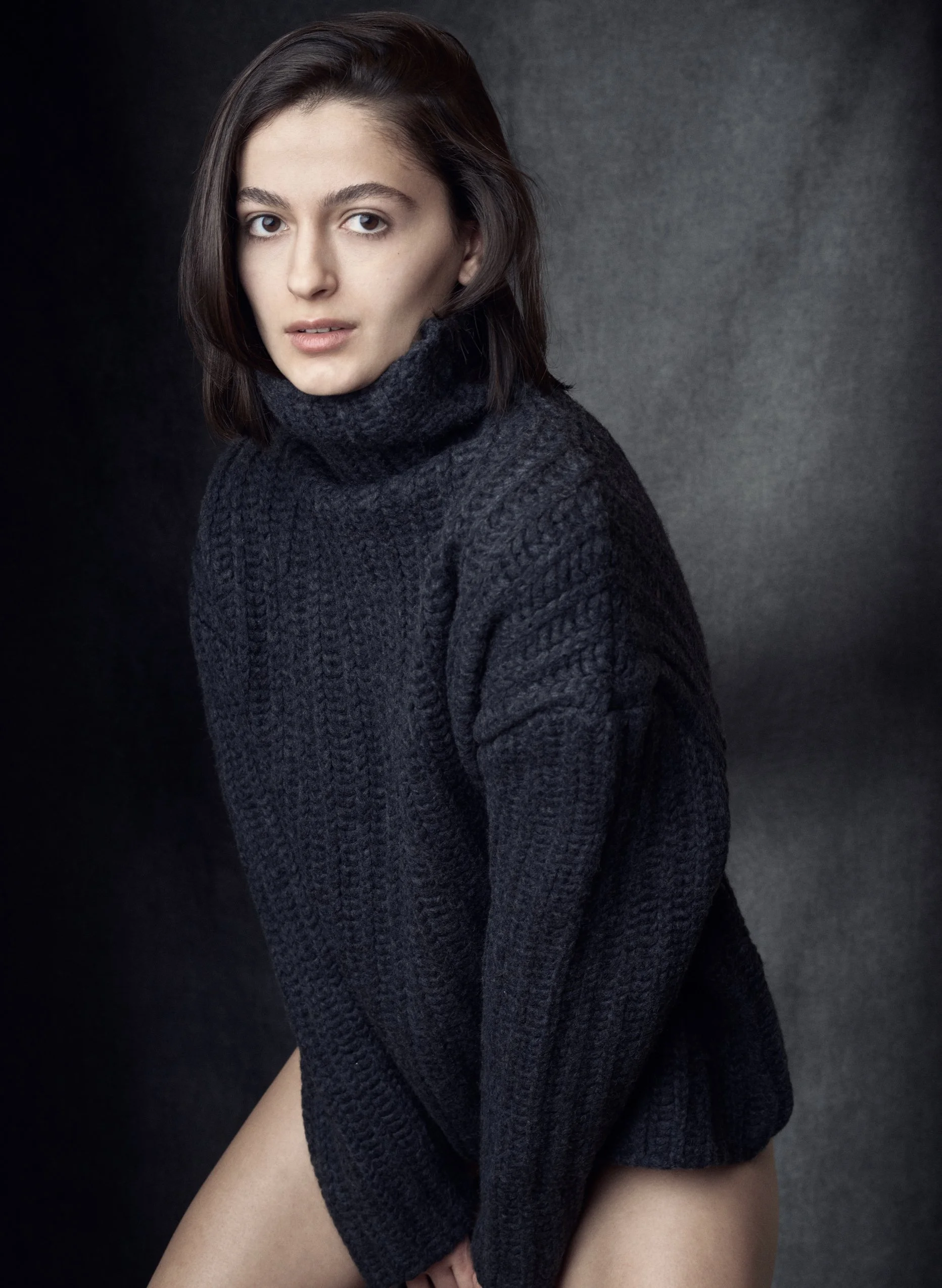 A young woman with short brown hair and light skin, wearing a dark knitted turtleneck sweater, sitting against a dark background.