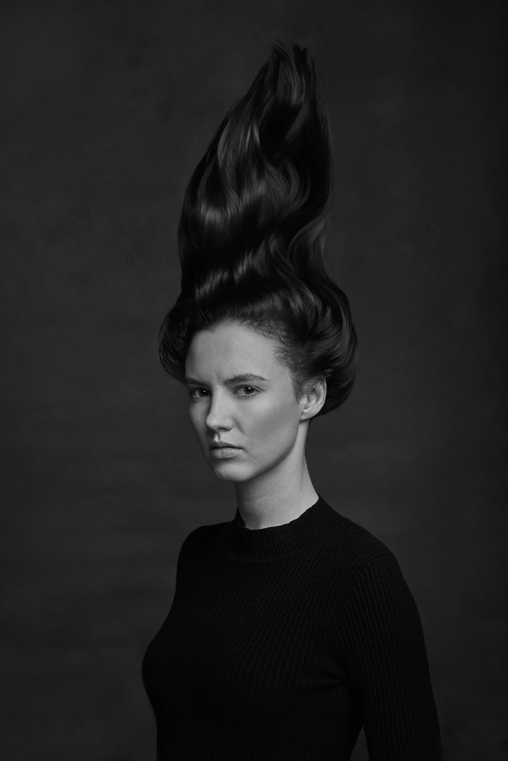 Black and white portrait of a woman with long, wavy hair styled upwards and a serious facial expression, wearing a black top.