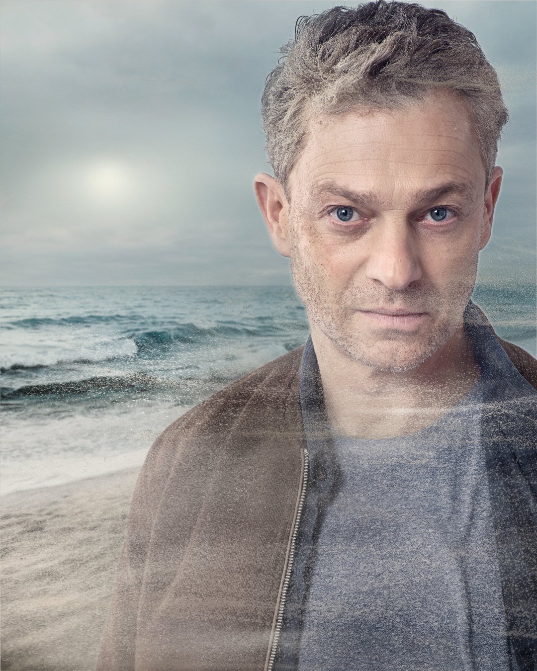 A man with gray hair and blue eyes looking at the camera, with a beach and ocean waves in the background, seen through a transparent surface.