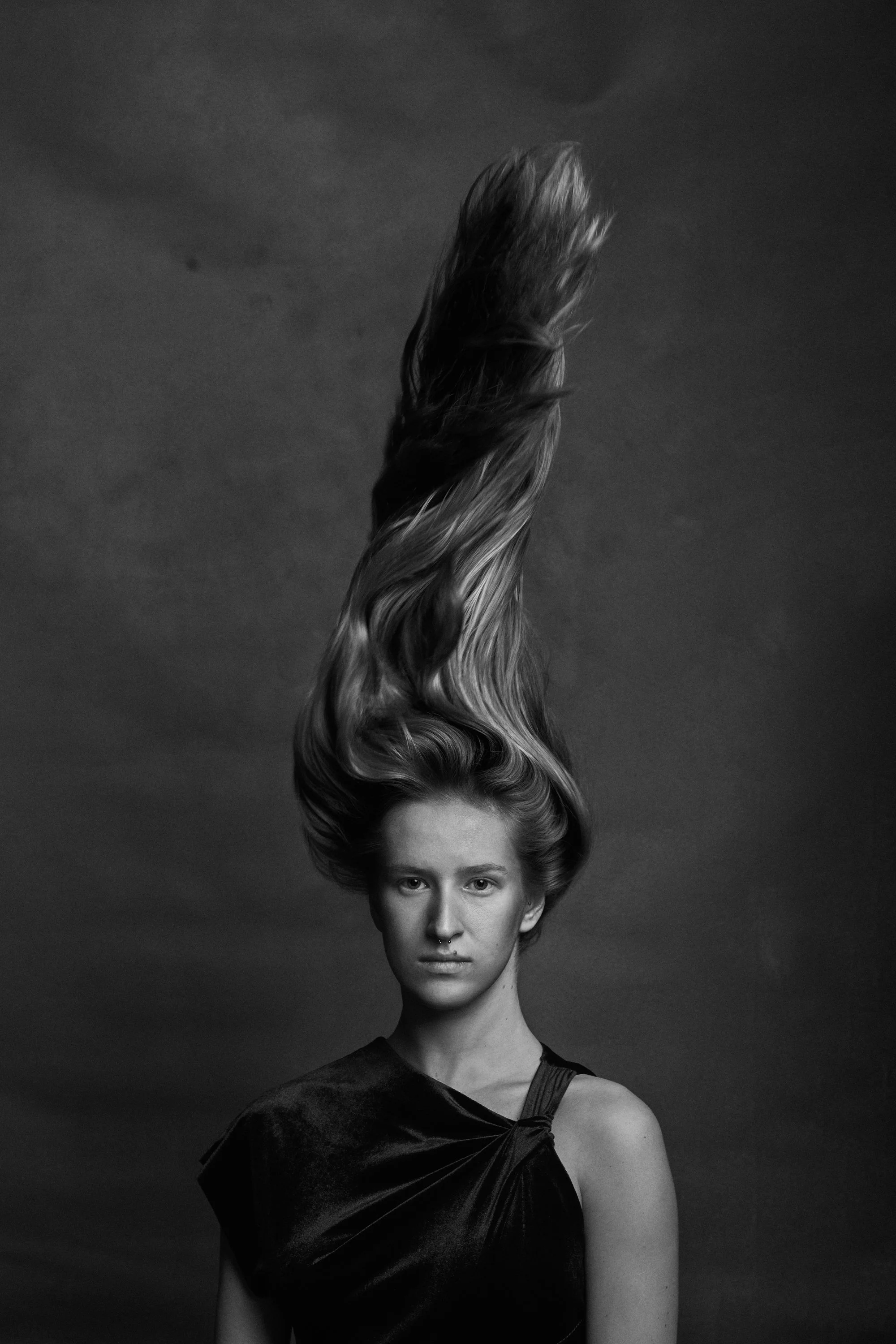 Black and white portrait of a woman with long, wavy hair flowing upward, wearing a dark sleeveless dress, looking directly at the camera against a plain dark background.