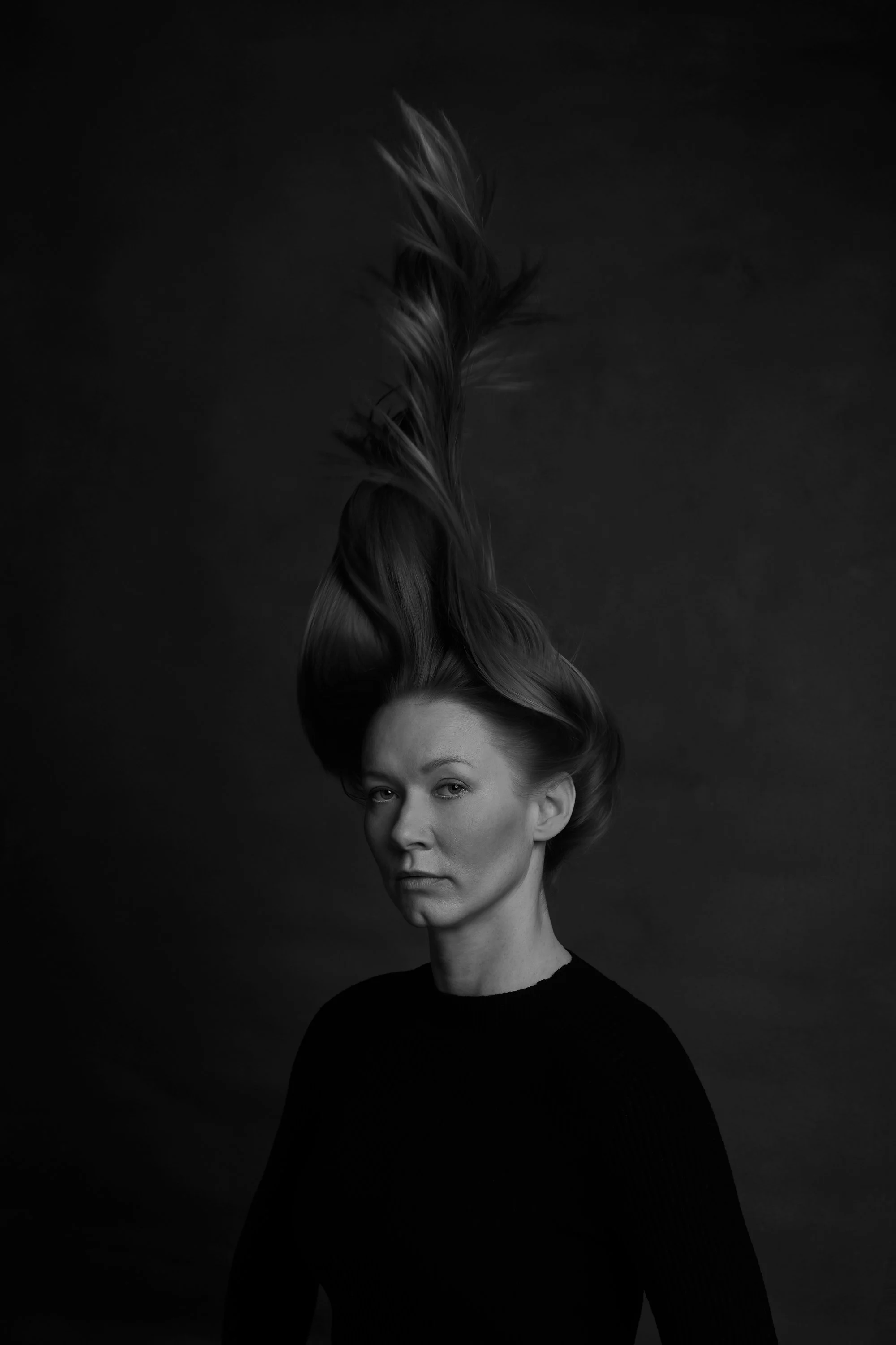 Black and white portrait of a woman with an elaborate updo hairstyle, looking directly at the camera against a dark background.