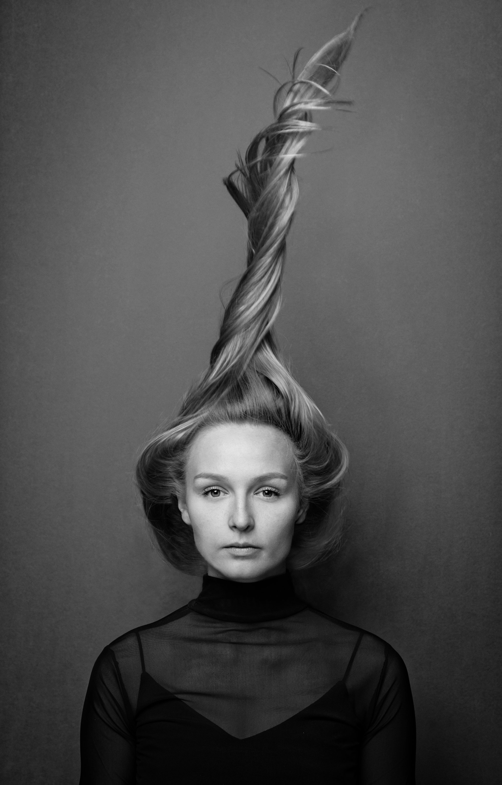 Black and white photo of a woman with an elegant hairstyle, her hair twisted upwards with curled ends, wearing a sheer long-sleeve top with a high neckline and subtle pattern.