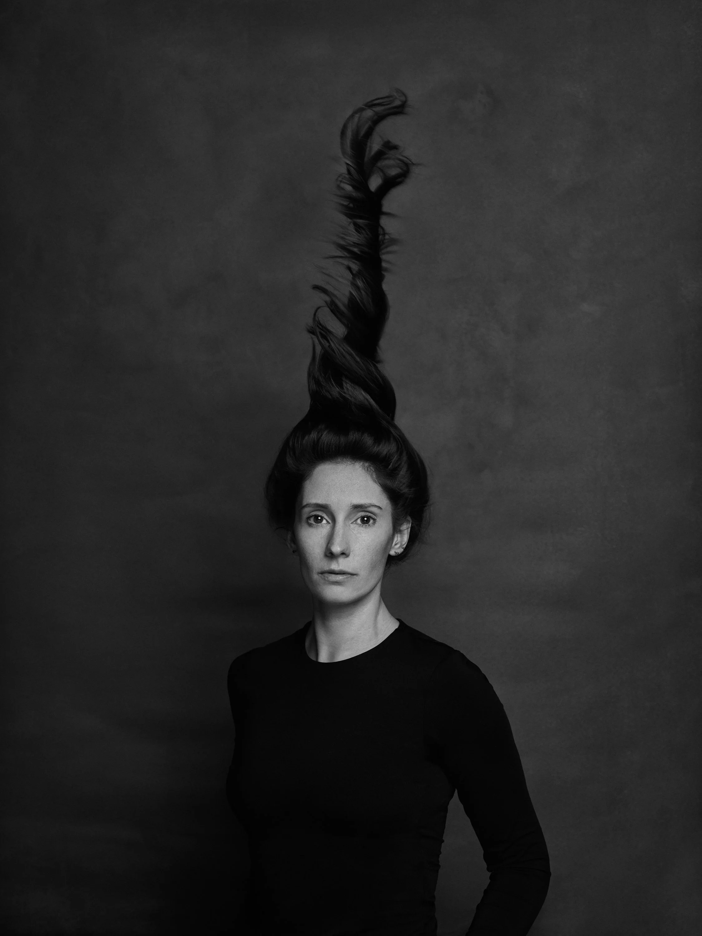 A black and white portrait of a woman with long hair styled in a high, twisted updo. She is wearing a black shirt and has a neutral expression.