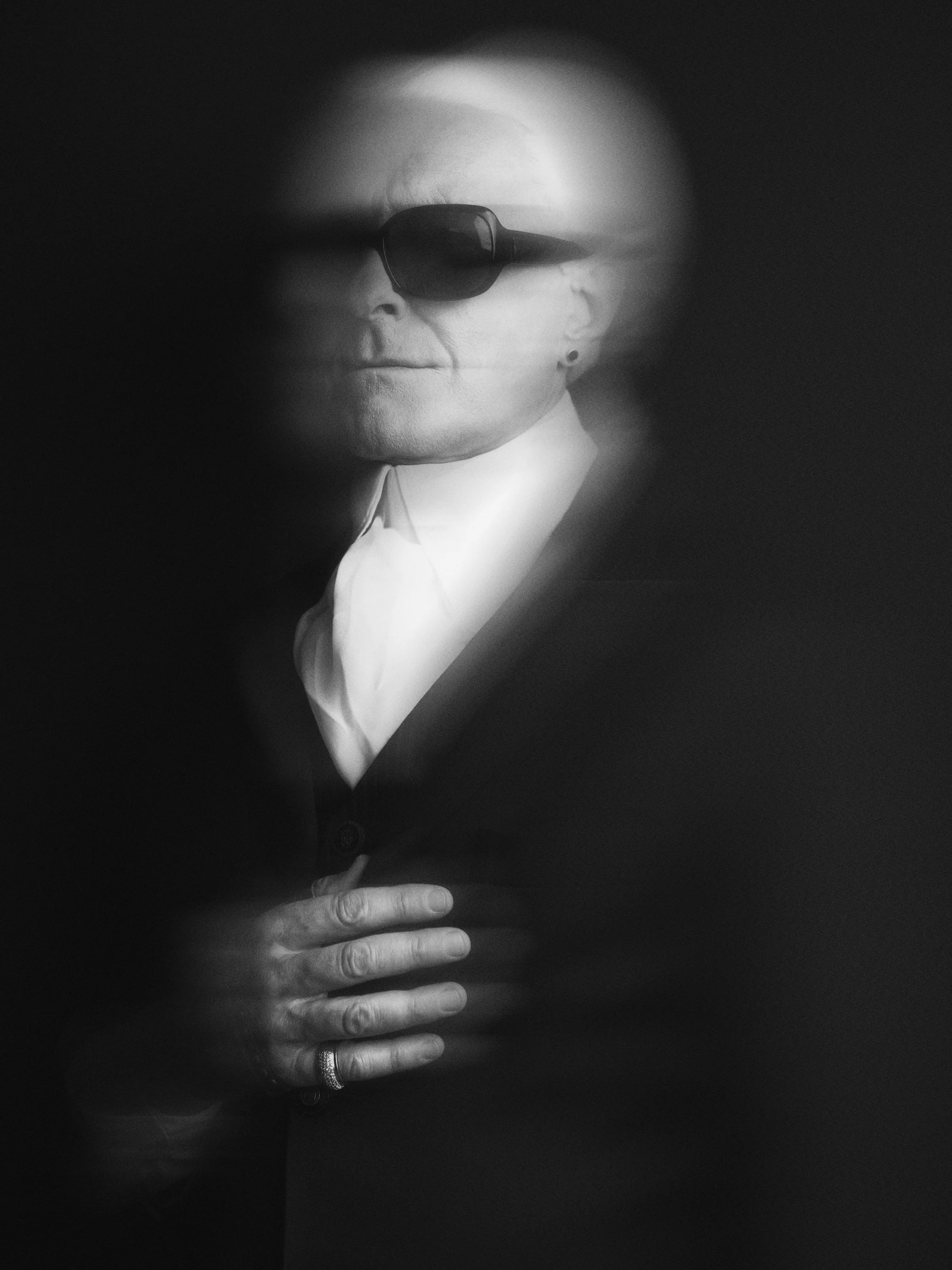 Black and white photo of an elderly woman wearing dark sunglasses, with one hand resting on her chest and the other partially visible. She has short hair and a small earring.