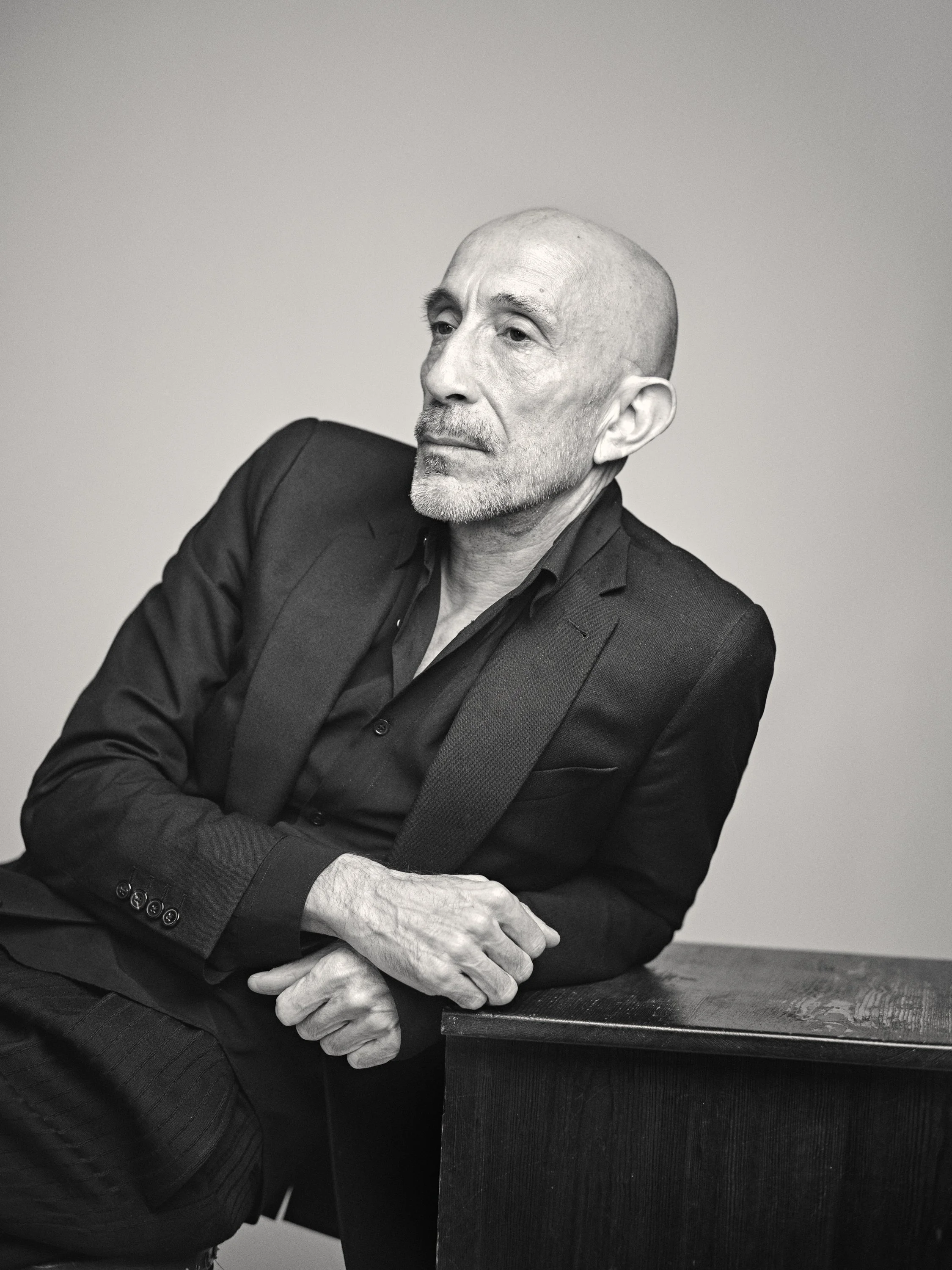 Black and white photo of a middle-aged man with a beard, wearing a dark blazer, sitting at a table with his arms crossed in front of him, looking contemplative.
