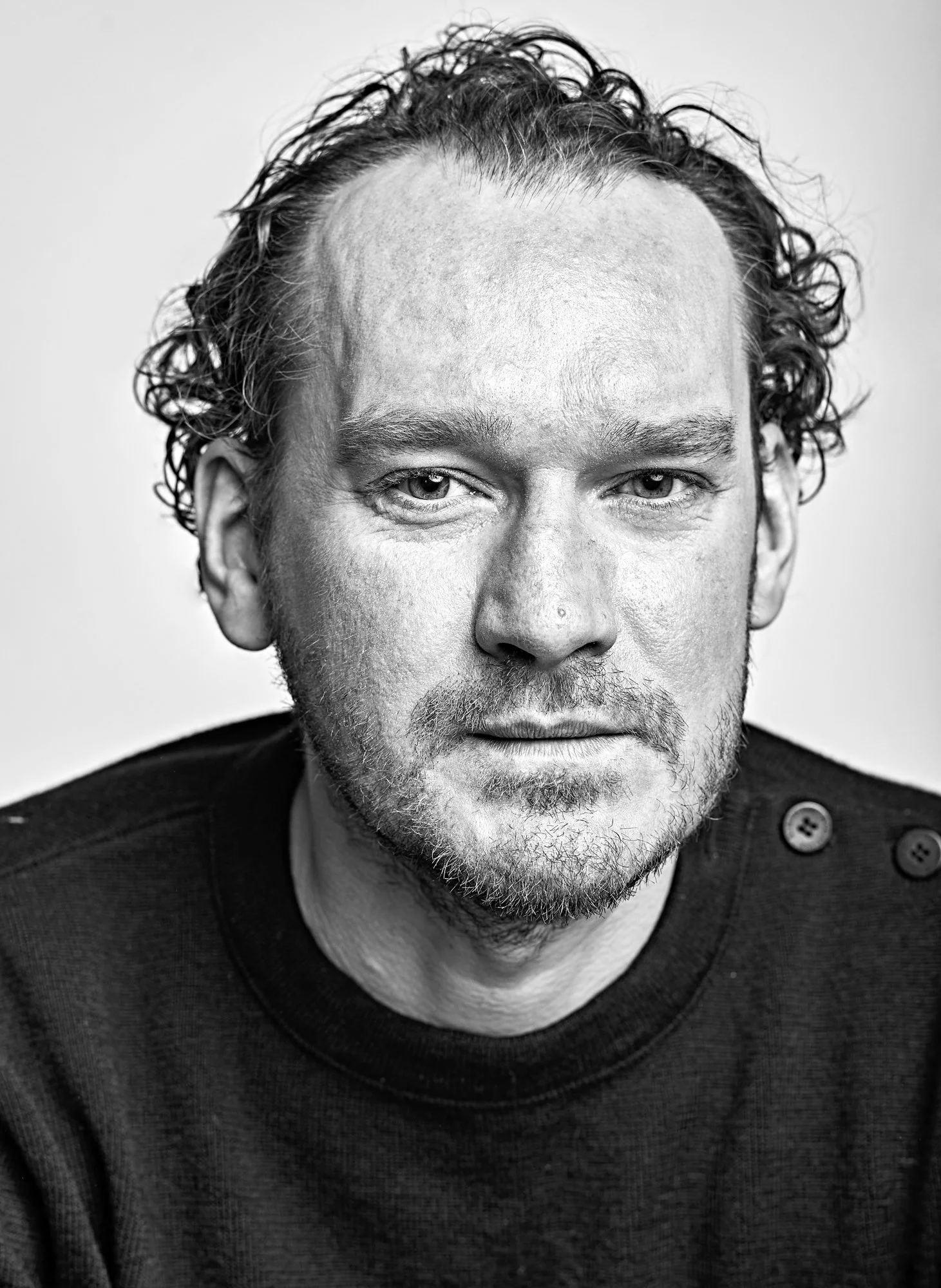 Black and white close-up photograph of a man with curly hair, blue eyes, and facial hair, wearing a dark sweater with buttons on the shoulder.