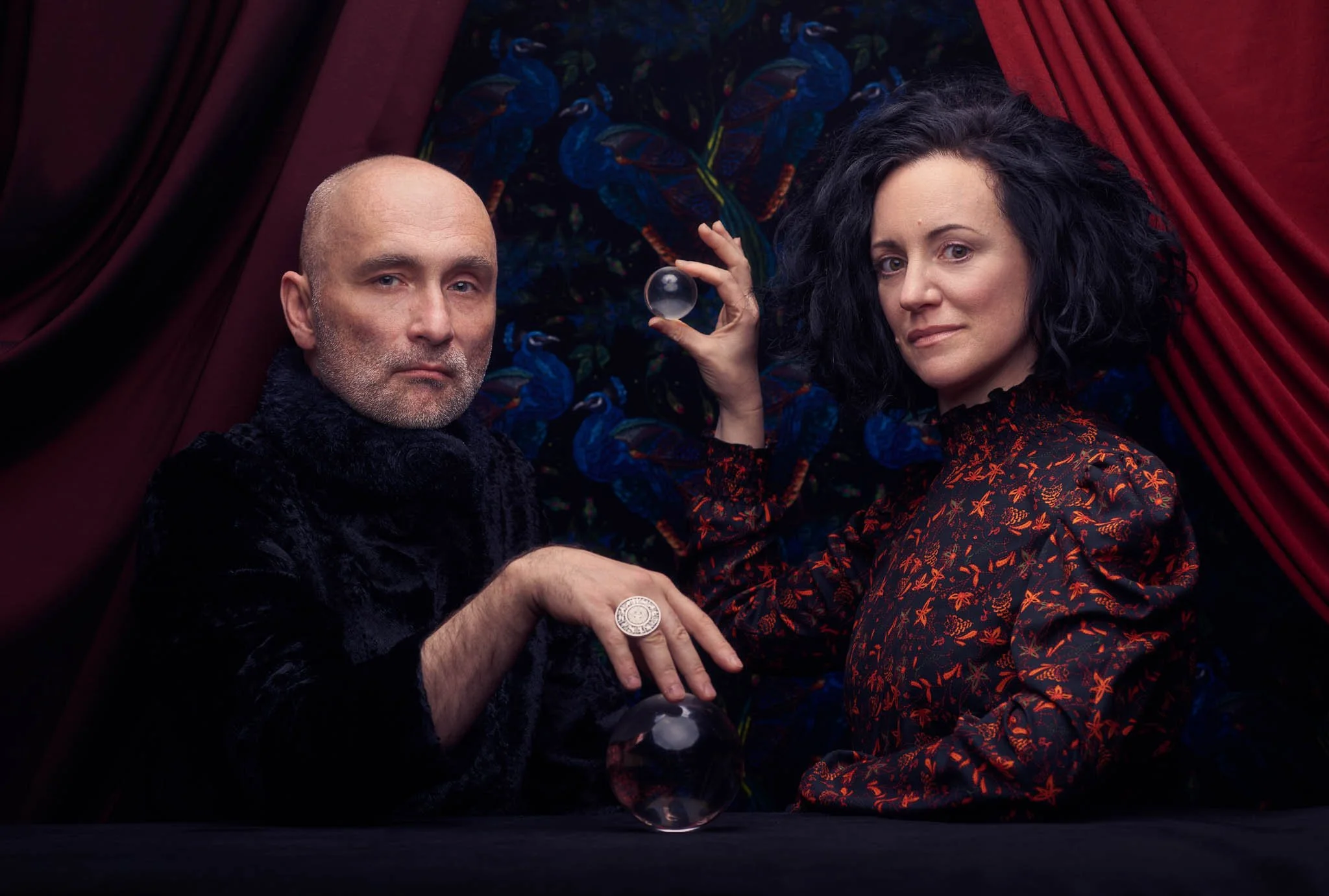 A man and woman are lying down on a dark surface in front of red curtains, with dark floral fabric behind them. The man has a bald head and gray beard, wearing a black furry coat and a large ring. The woman has black curly hair, wearing a black and orange patterned dress, and is holding a glass sphere.