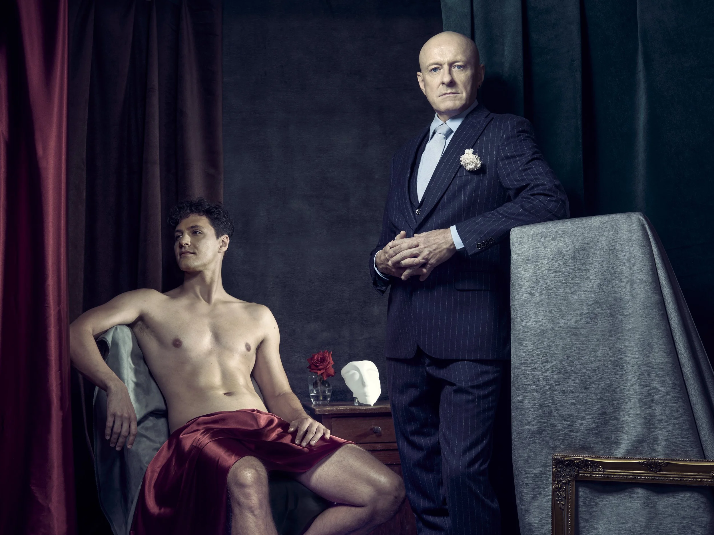 A shirtless man sitting on a bed with red sheets, a woman’s hand resting on his thighs, and a statue of a head and a flower on a table beside him, with a man in a pinstripe suit standing nearby, in a room with dark curtains and a dark wall.