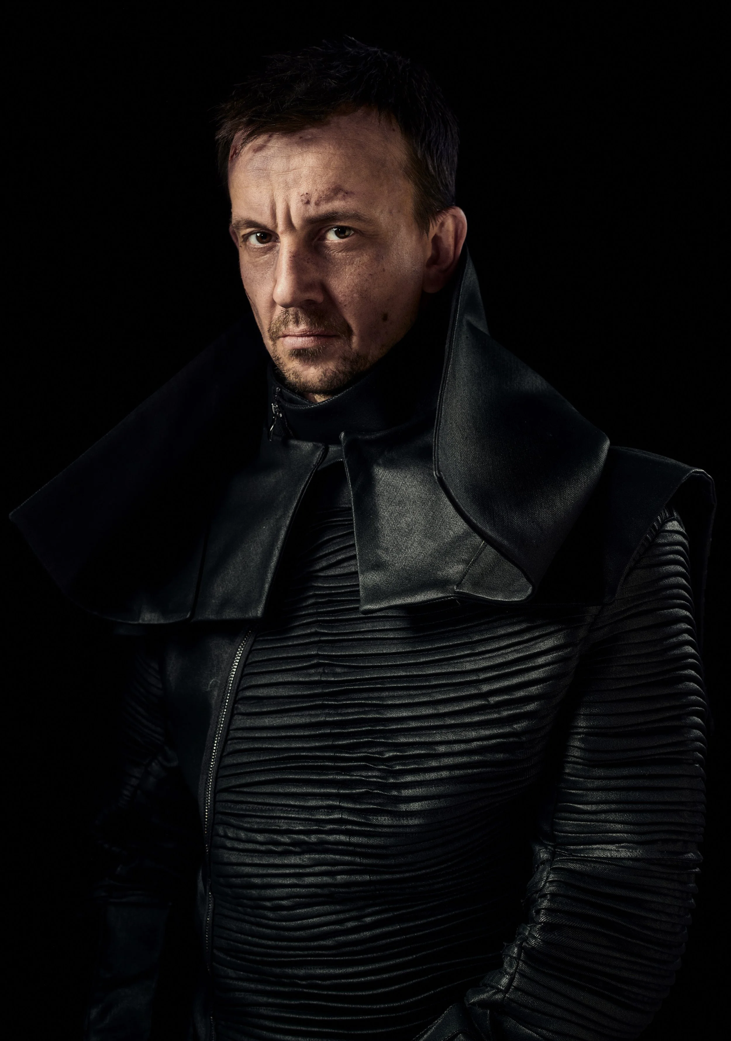 A man with short dark hair, wearing a black leather and ribbed jacket, looking directly at the camera against a dark background.