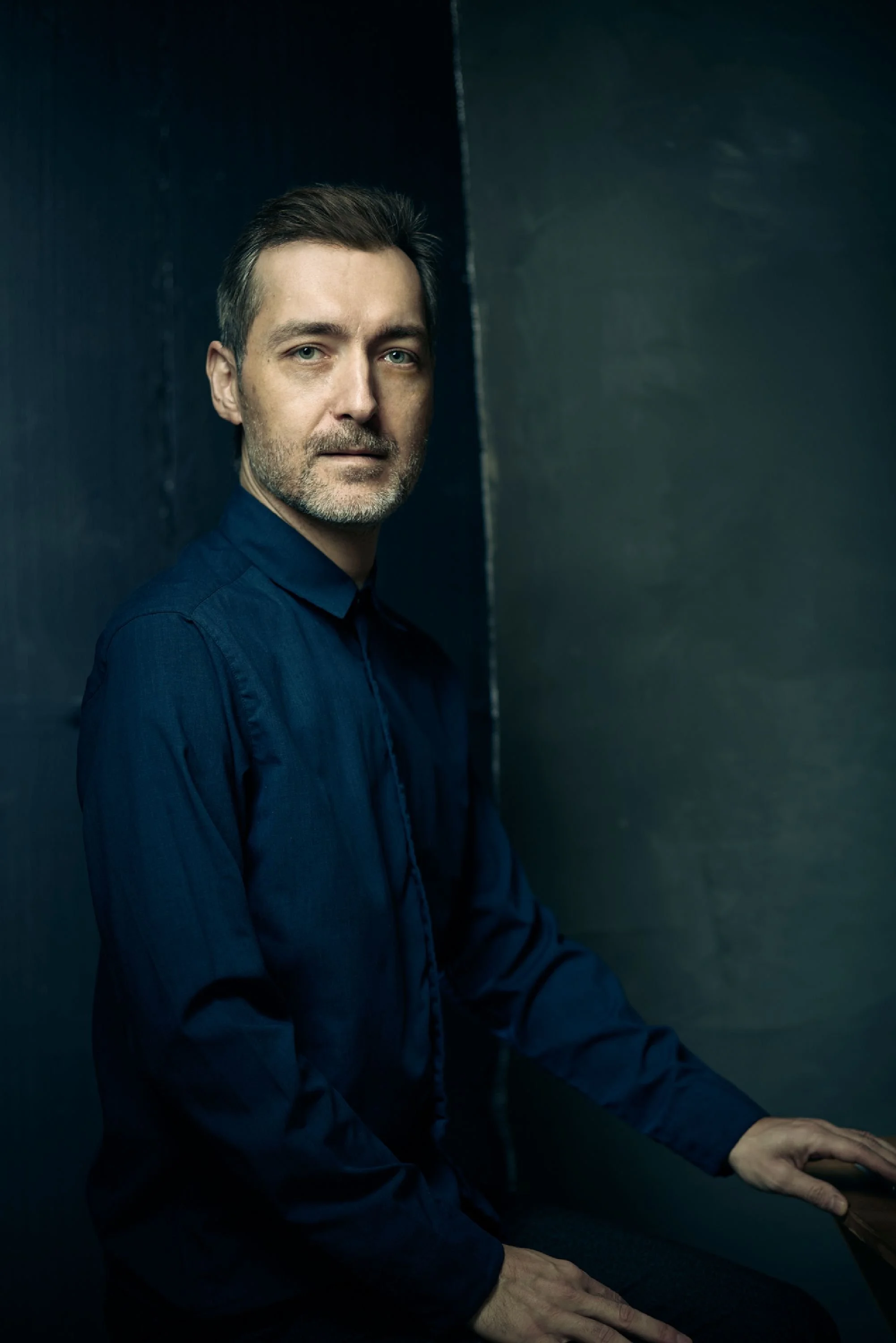 A man with short dark hair, beard, and mustache, wearing a dark blue button-up shirt, seated against a two-tone dark background, with one hand resting on his knee and the other hand's fingers visible at the bottom edge.