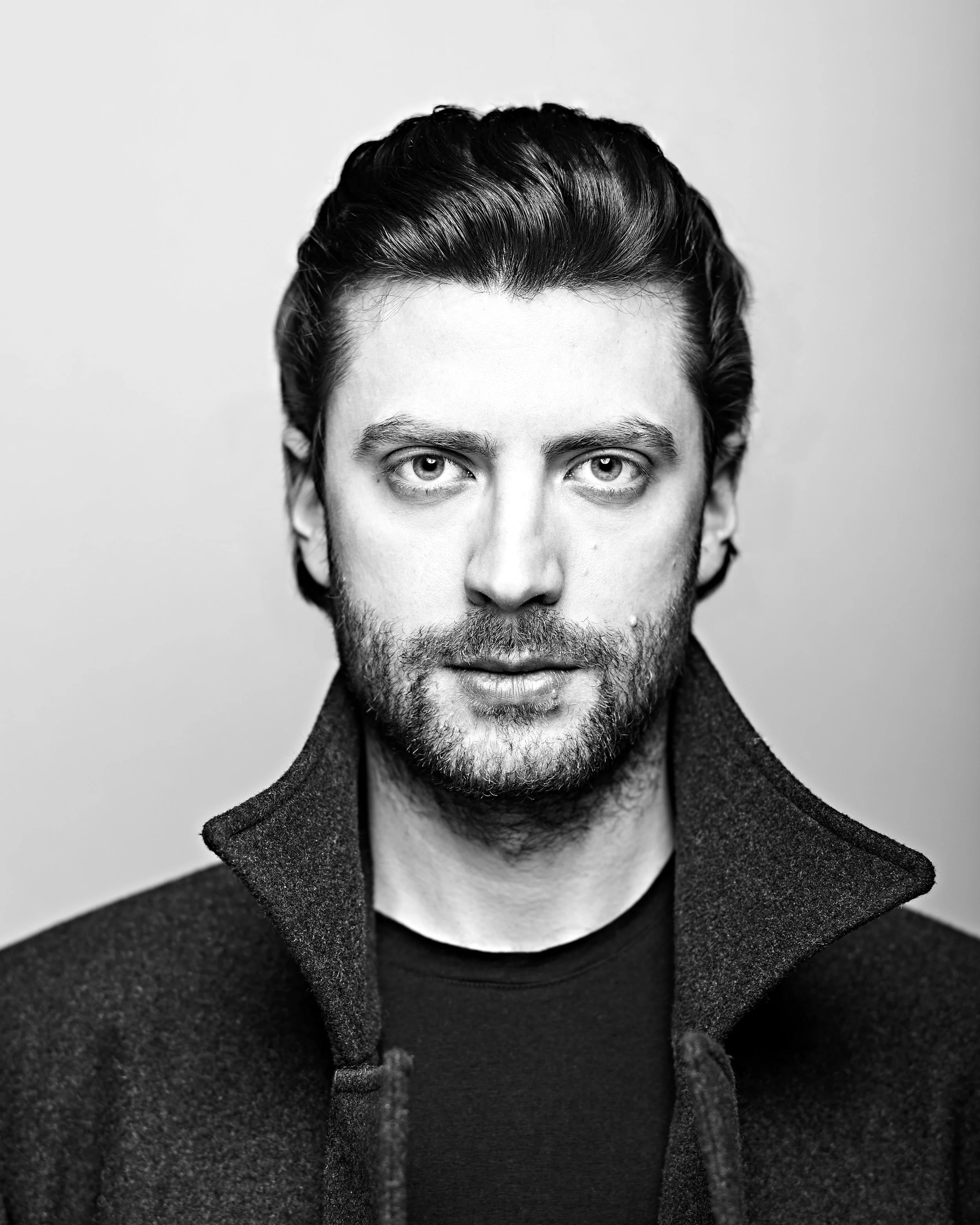 Close-up of a man's face with intense expression, dark hair styled back, and a beard, wearing a dark coat, black shirt, against a plain background.